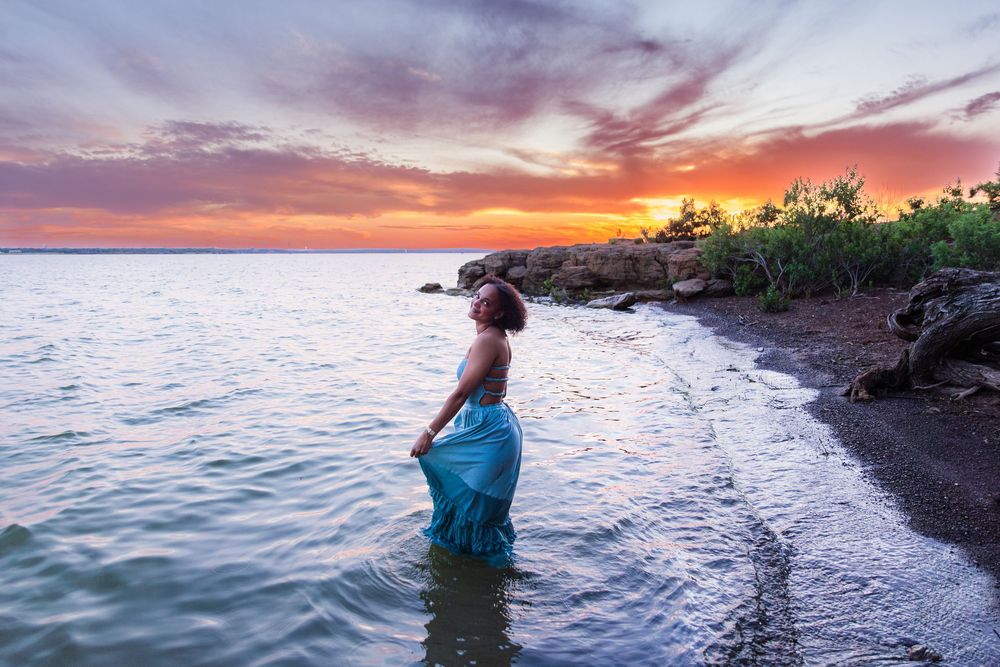 Woman in blue skirt standing in ocean water at sunset with vibrant orange and purple sky.