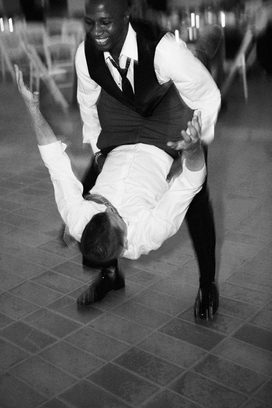 Two men in formal attire perform an energetic dance move on a tiled floor in this black and white photo.
