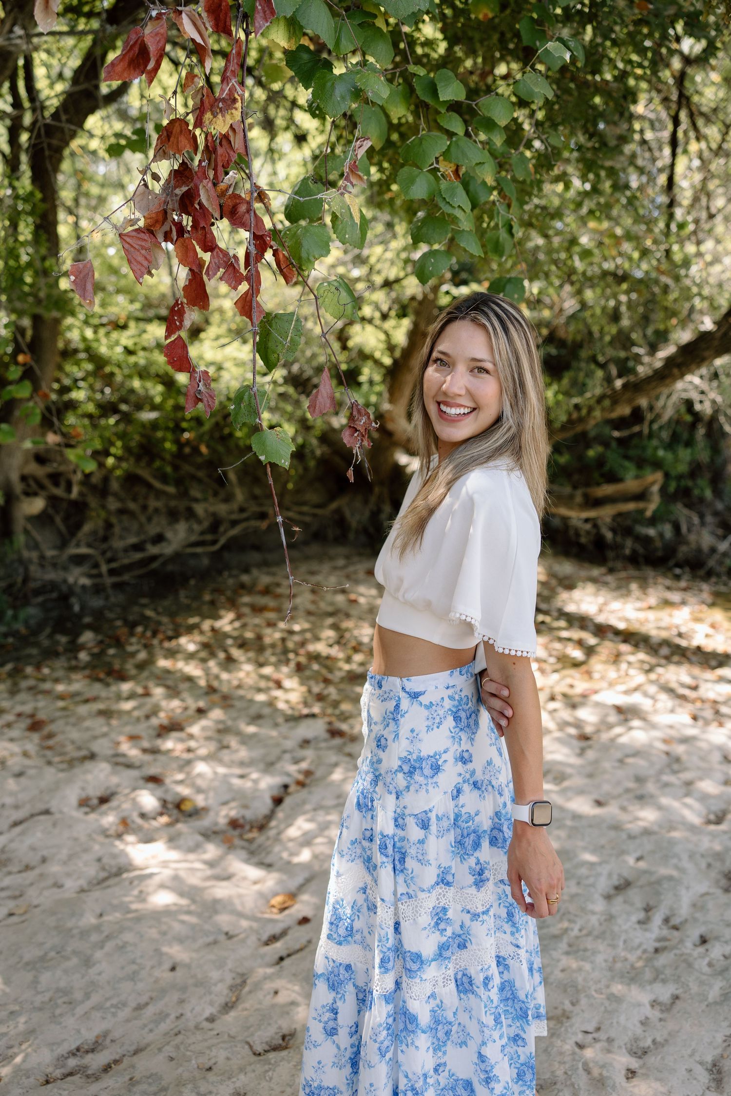 Smiling woman in white crop top and blue floral maxi skirt posing outdoors near lush green trees on sandy ground.