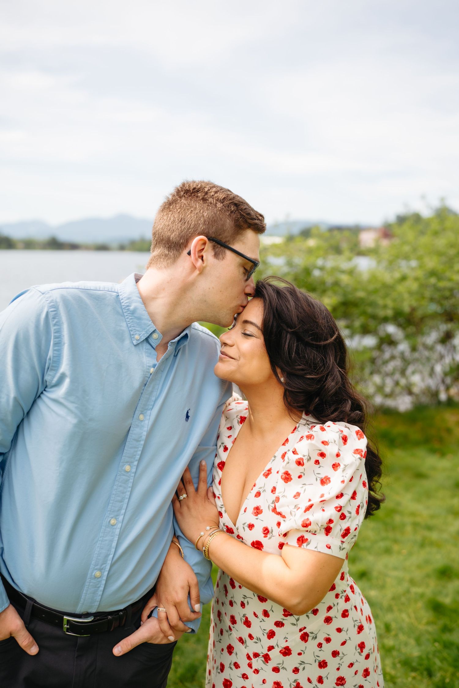 Couple sharing tender forehead kiss outdoors near lake, man in blue shirt, woman in red floral dress.