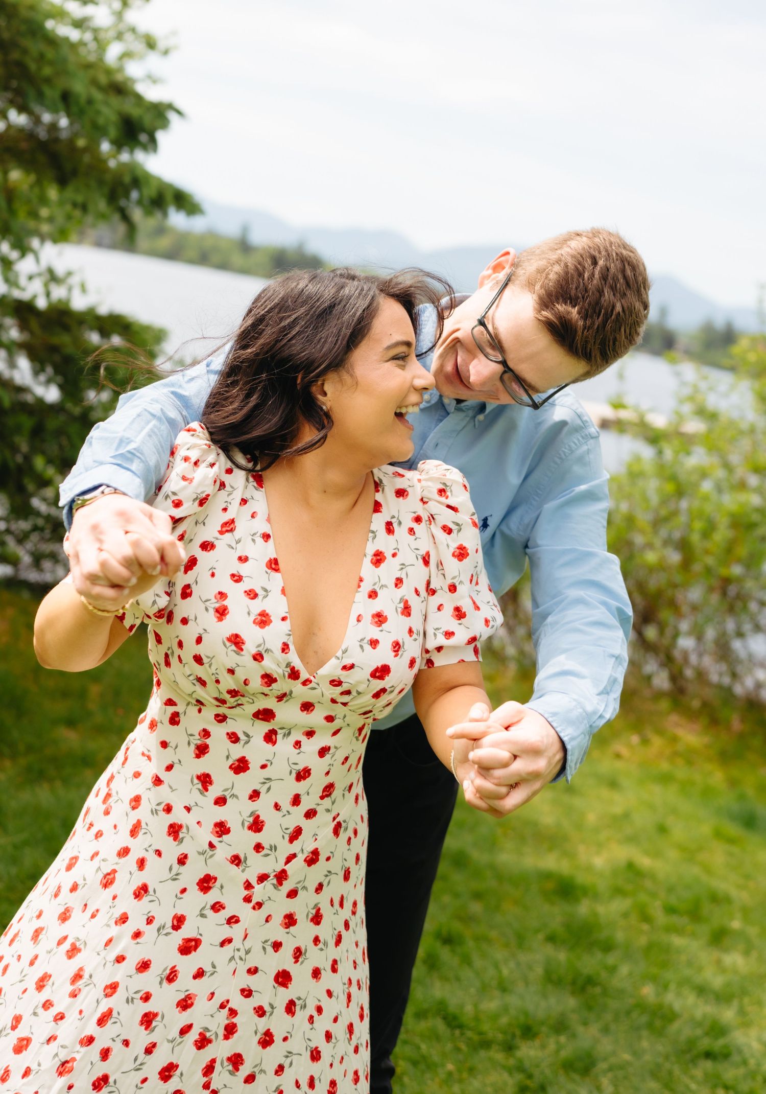 Happy couple dancing and laughing together near a lake, woman in red floral dress, man in blue shirt.