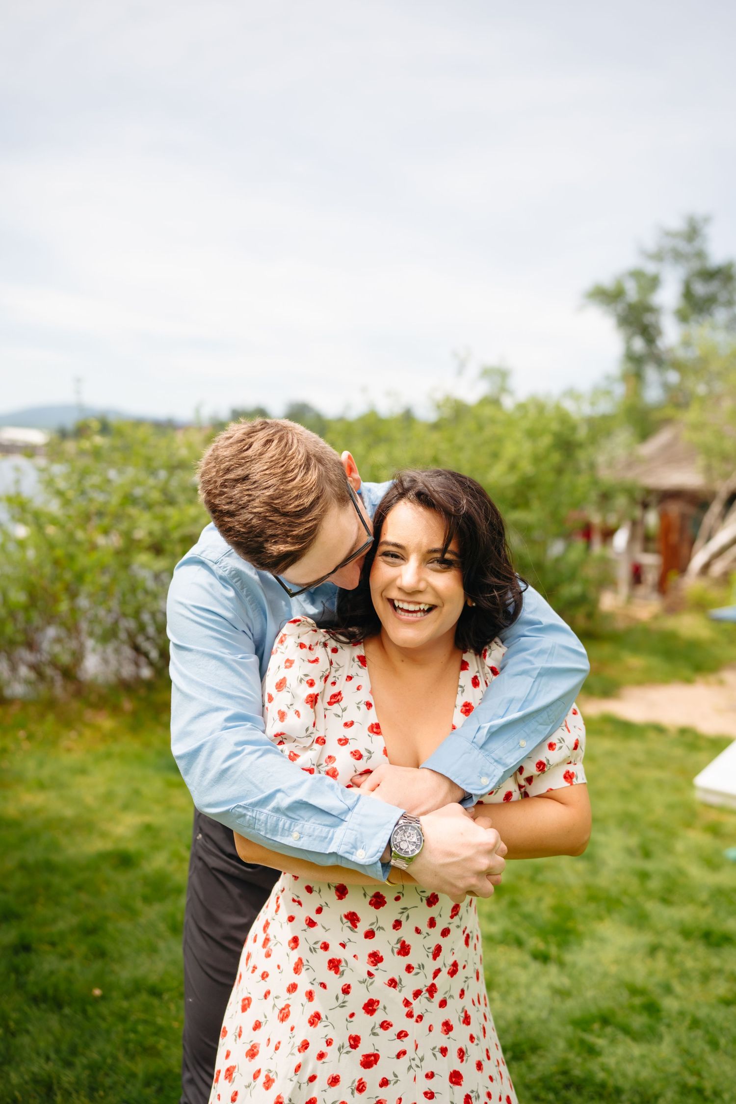 Man in blue shirt hugging woman in floral dress from behind in a green garden setting.