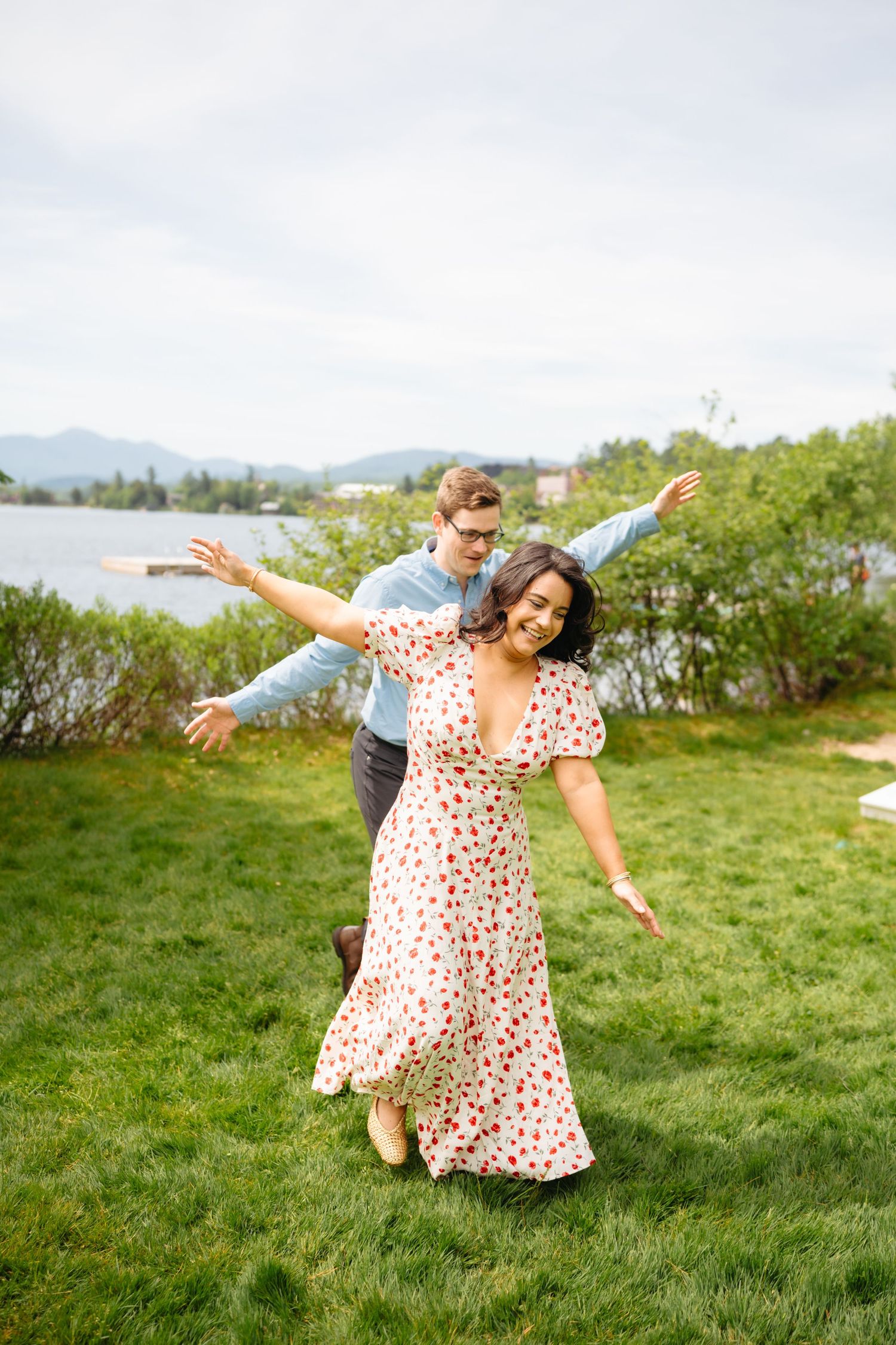 Joyful couple dancing on green lawn near a lake, woman in floral maxi dress with arms outstretched, man behind her smiling.