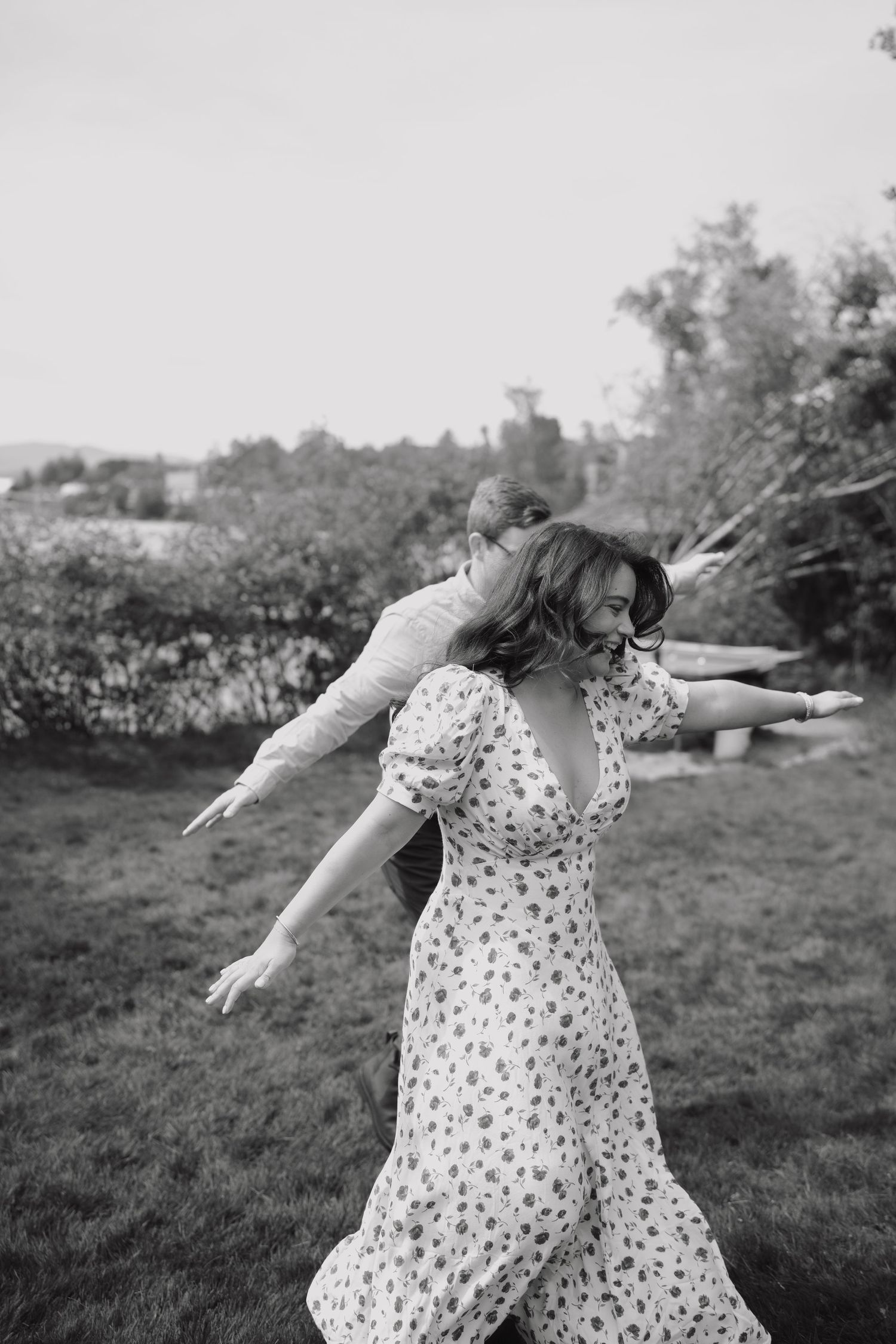 Black and white photo of a woman in a floral dress with arms outstretched, joyfully spinning outdoors with a child.