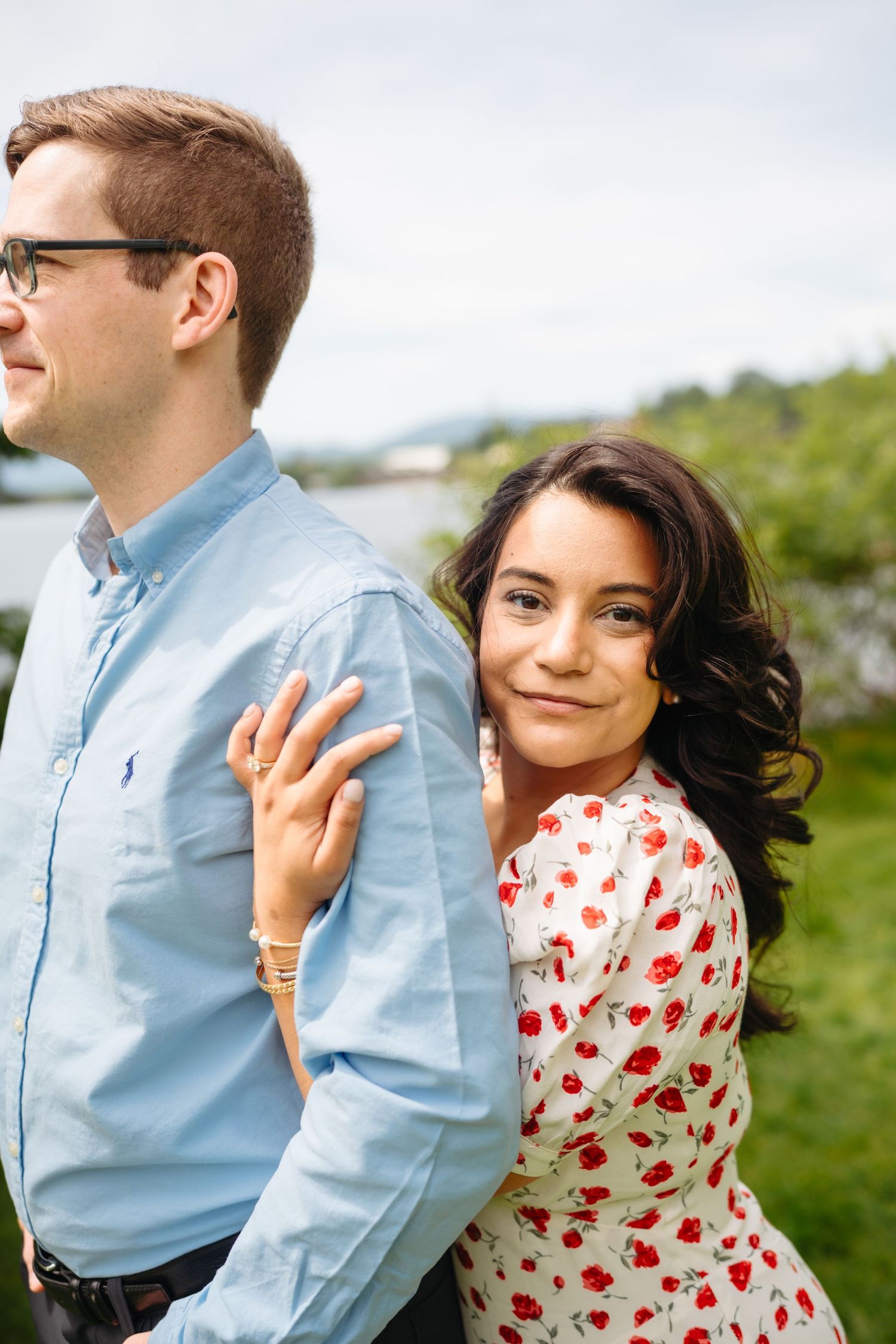 Engaged woman showing ring while leaning on man's back, smiling at camera near lake.