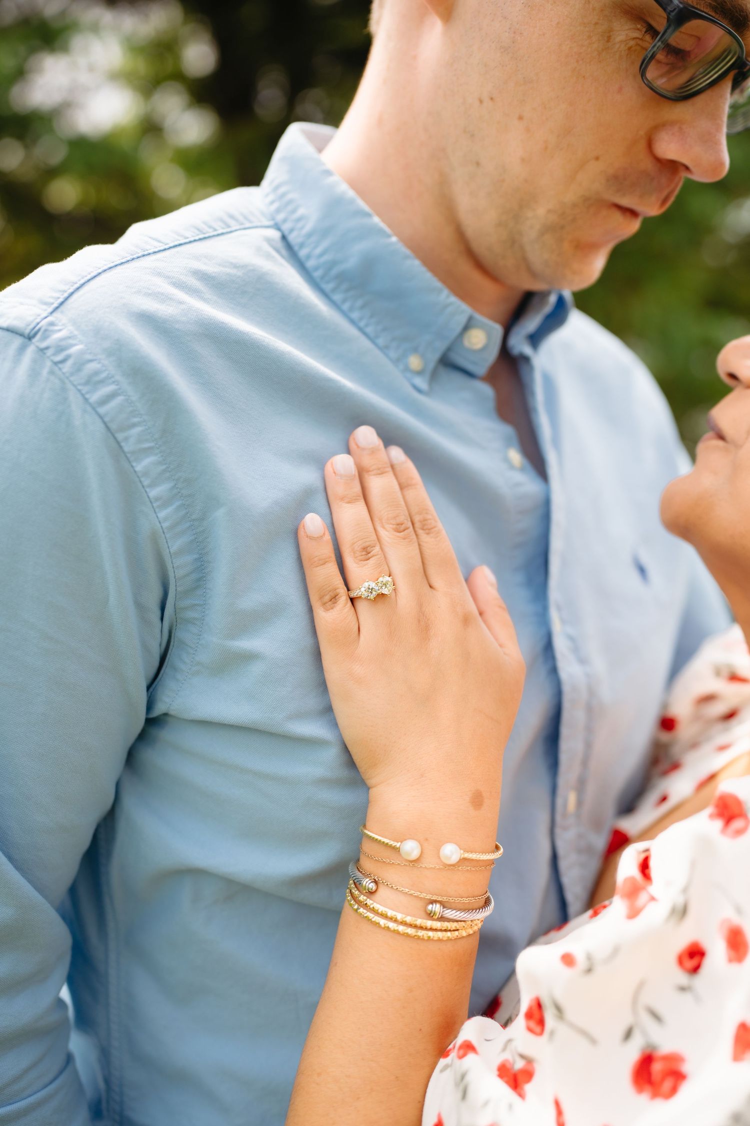 Close-up of a woman's hand with a gold engagement ring and bracelets resting on a man's light blue button-up shirt.