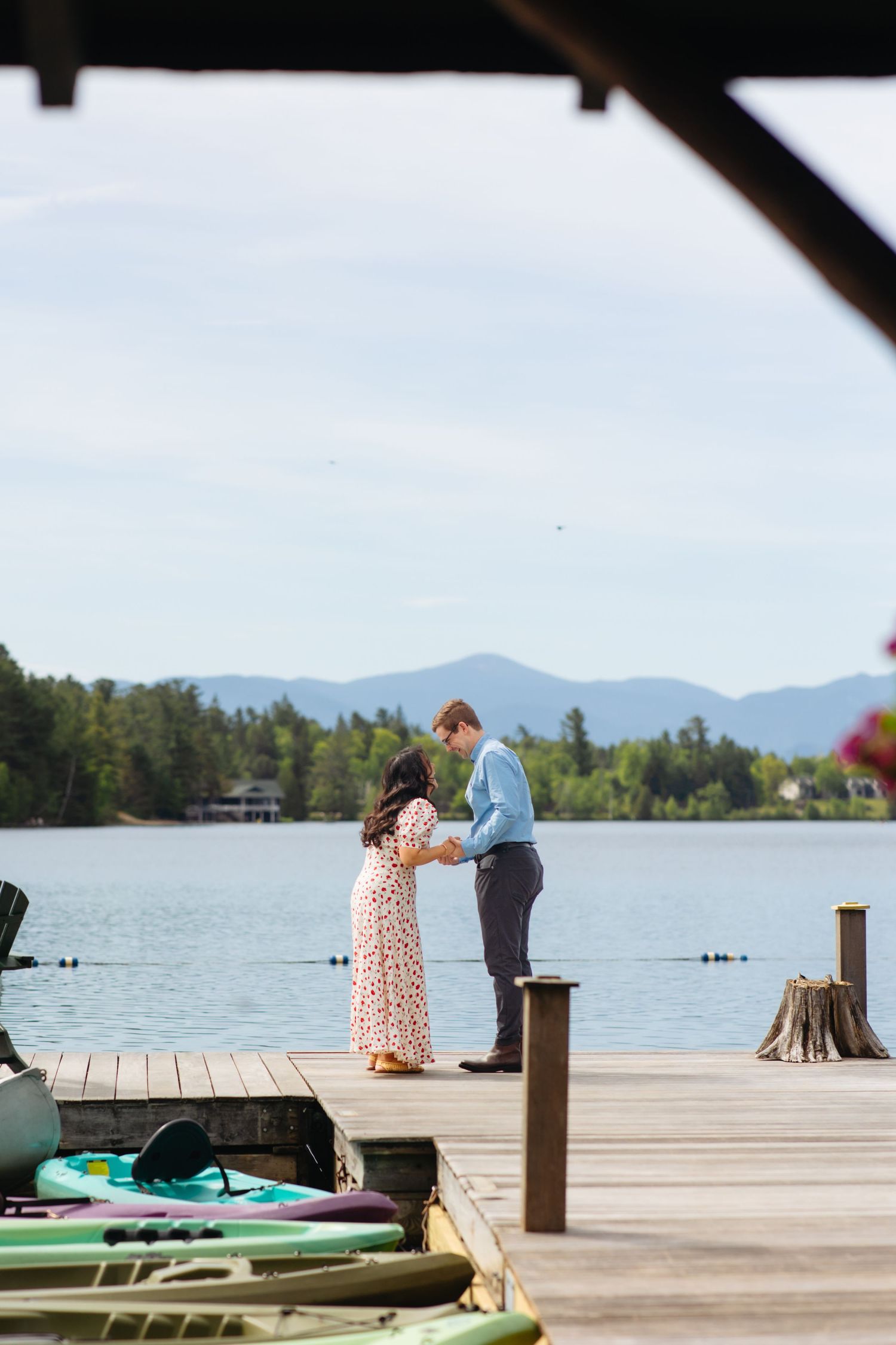 Couple holding hands on a lakeside dock with mountain views, kayaks nearby, on a sunny day.