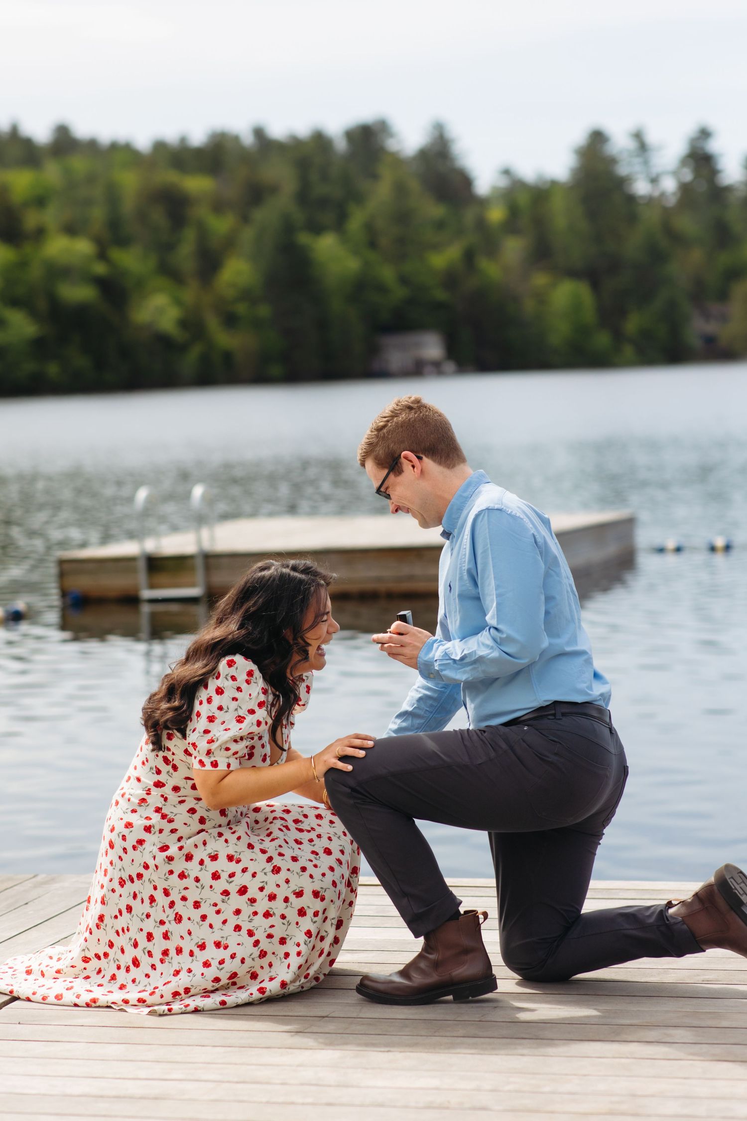 Man on one knee proposing to woman on wooden dock by scenic lake with forest backdrop.