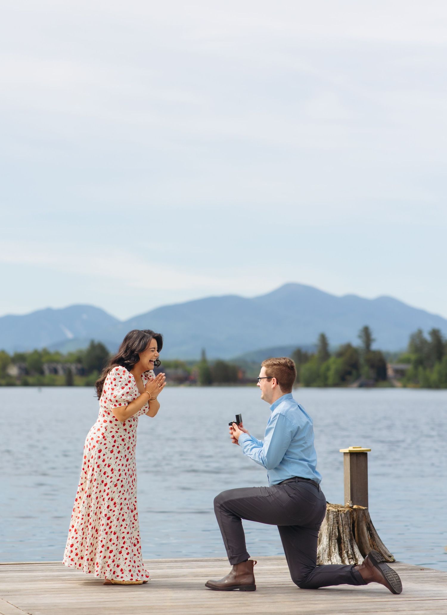 Man proposing on one knee to woman on a lakeside dock with mountains in background.