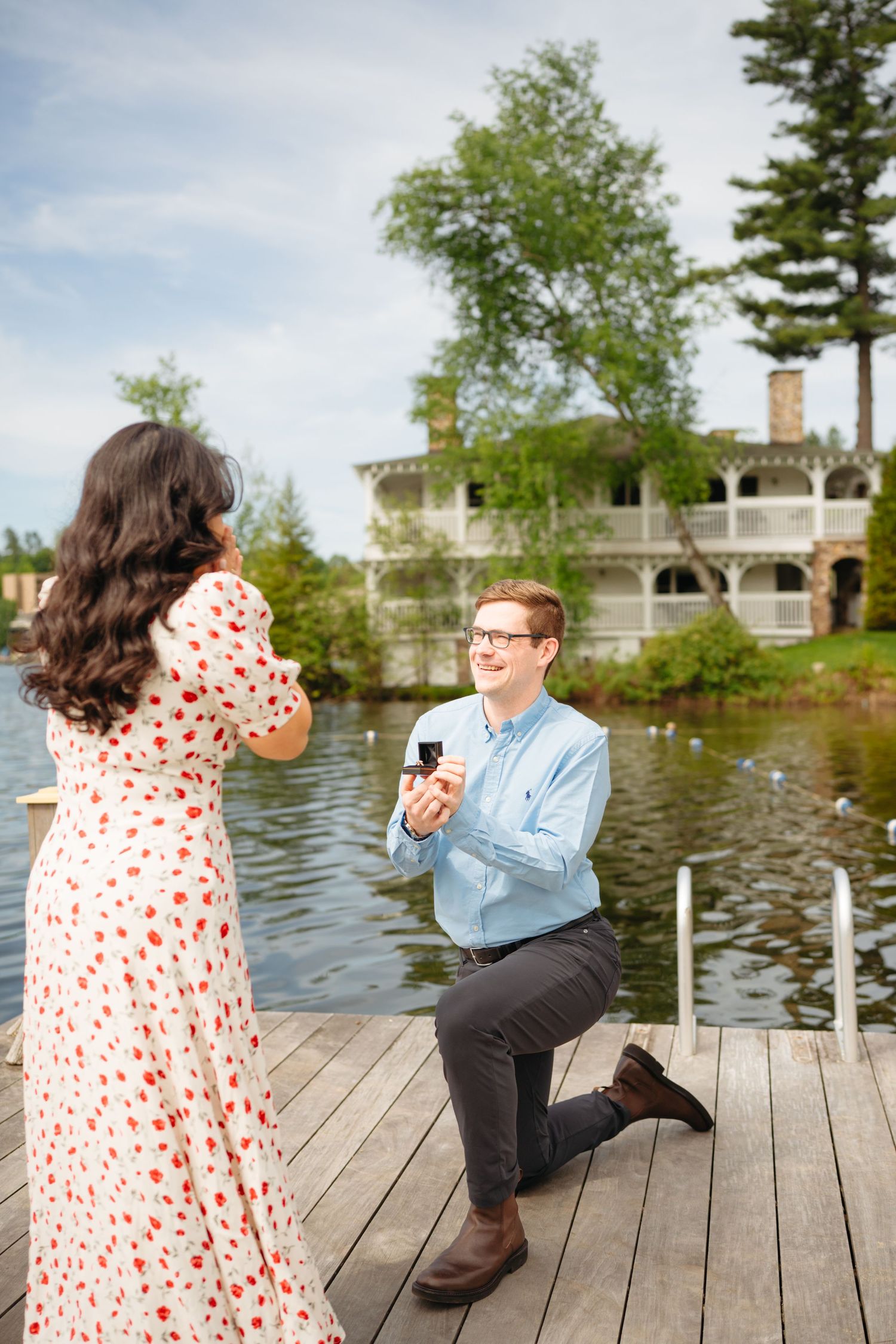 Man kneeling on a dock proposing to a woman in a floral dress by a lakeside mansion.