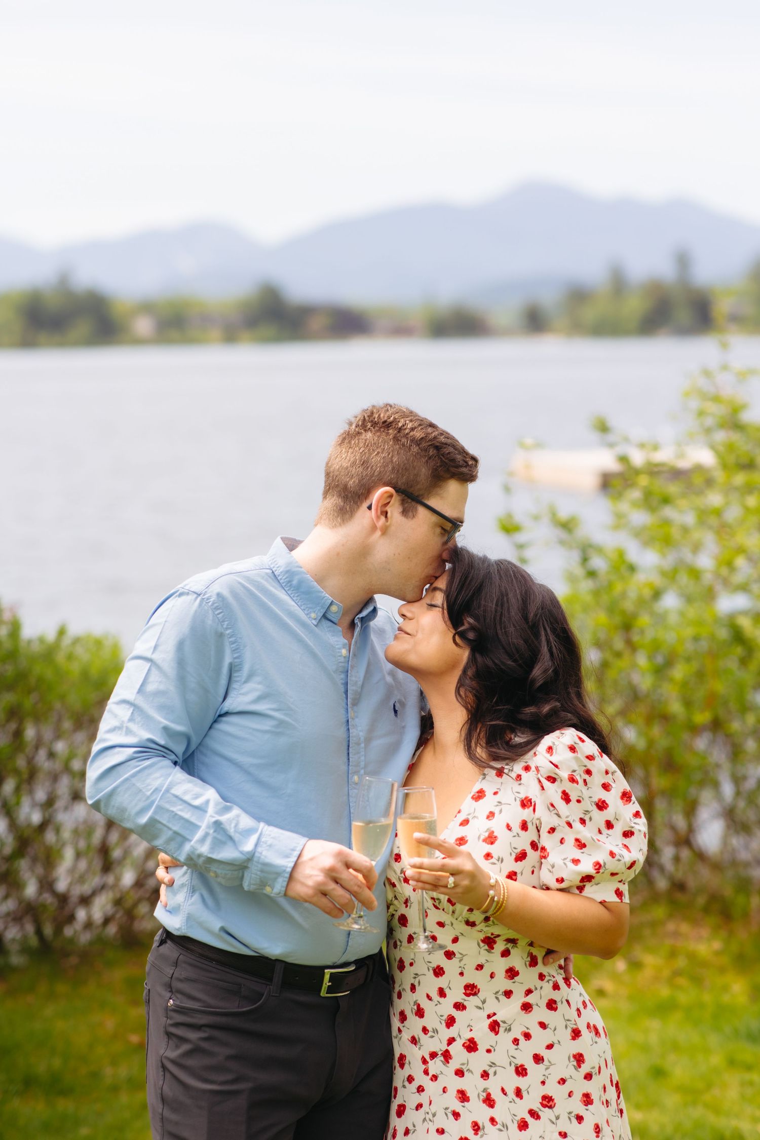 Newly engaged couple kissing while holding champagne glasses near a scenic mountain lake.