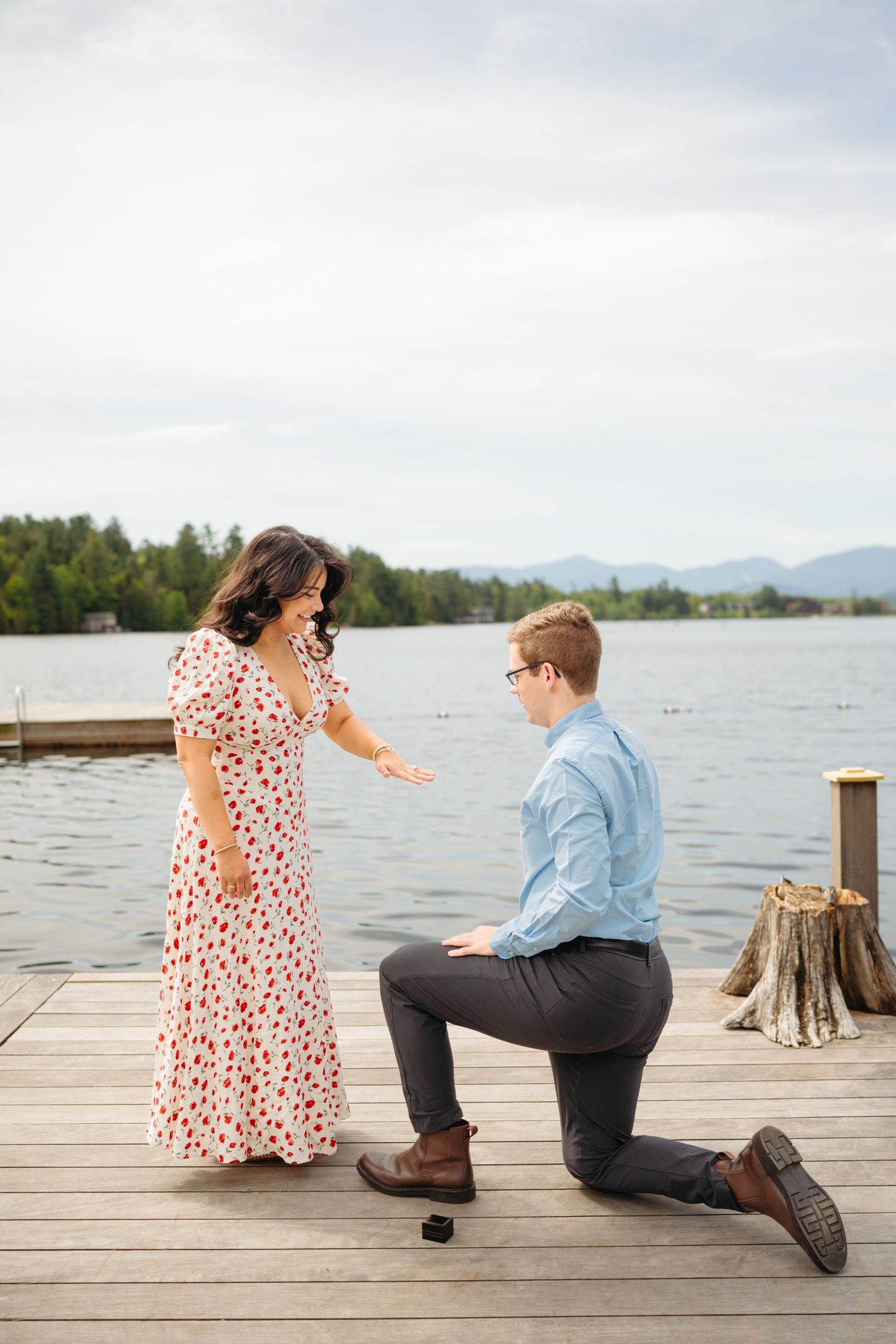 Man proposes on a lakeside dock, kneeling before a woman in a floral dress on a cloudy day.