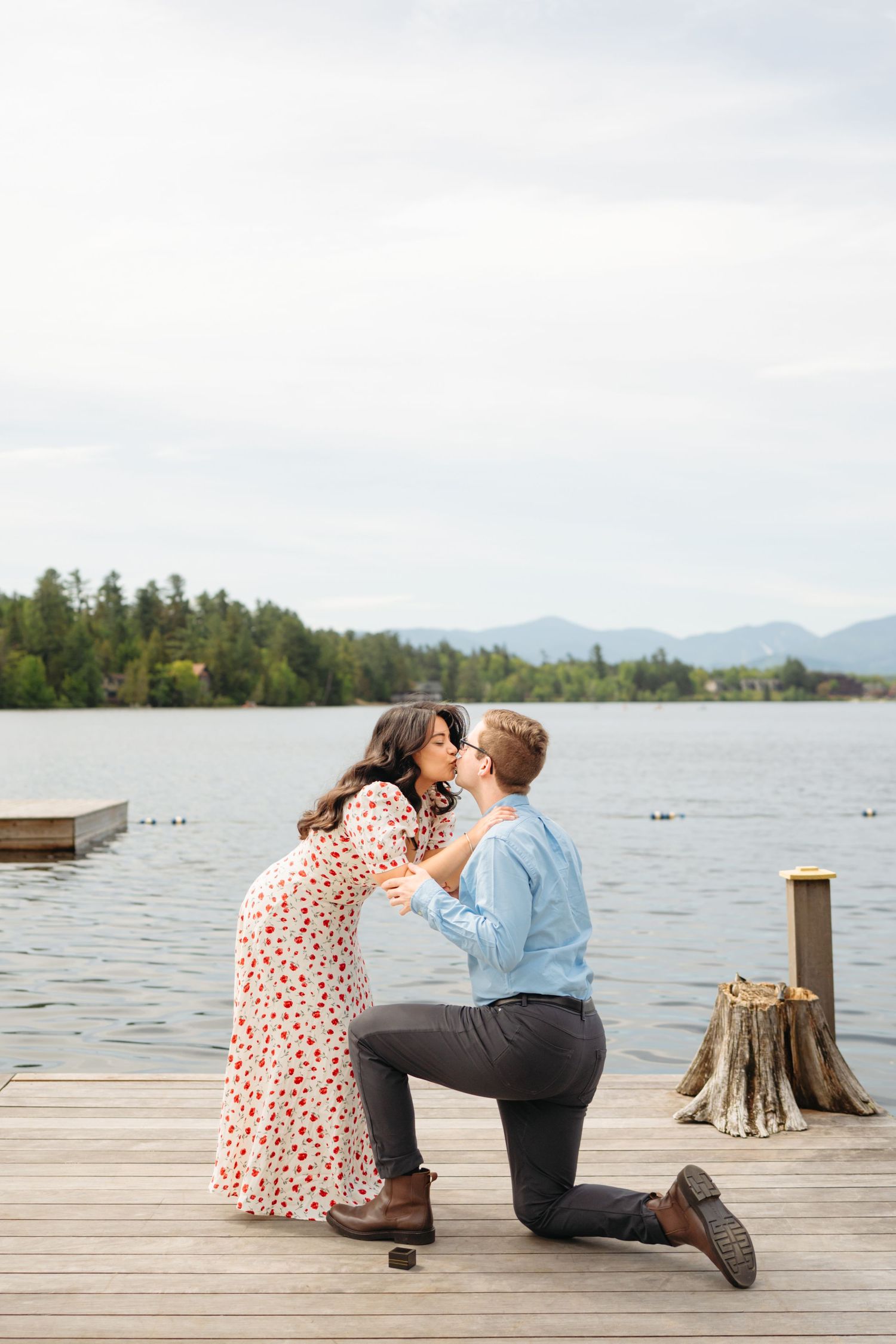 Person proposing on one knee to partner on a wooden dock by a scenic lake with mountains and trees in the background.