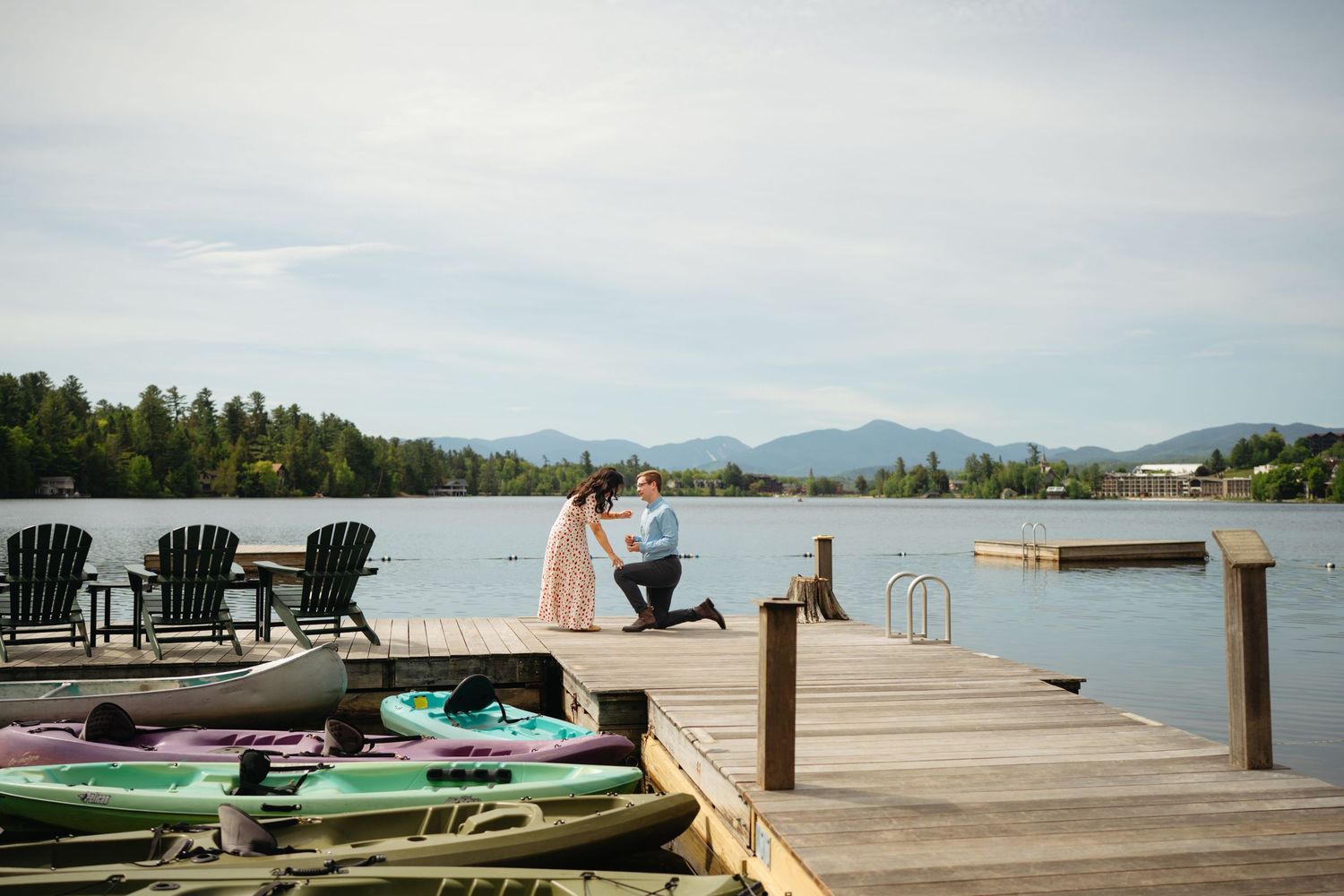 Man proposes on a wooden dock by a lake with kayaks and Adirondack chairs, mountains in the background.