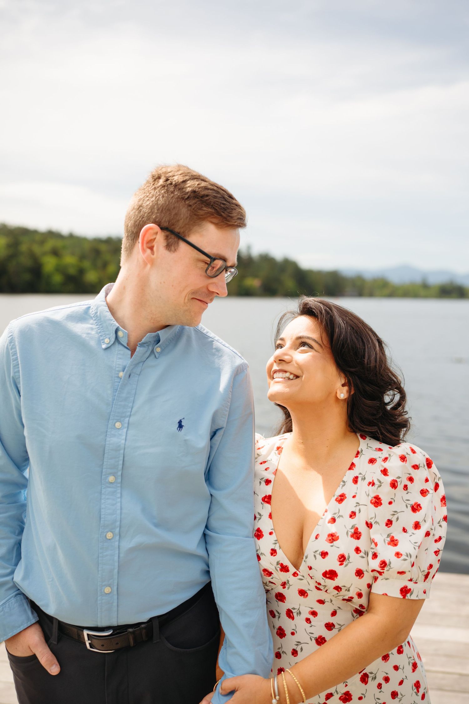 A smiling couple shares a tender moment on a lakeside dock, woman in floral dress, man in blue shirt.