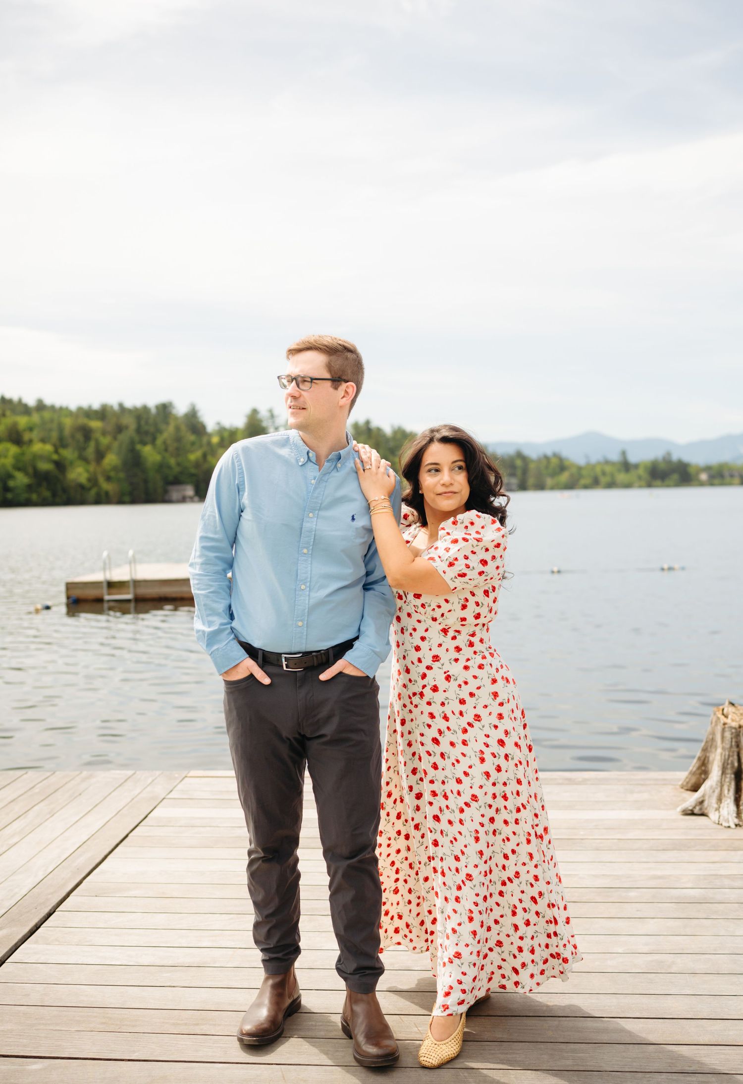 Couple posing on a lakeside dock, man in blue shirt and woman in floral dress, with mountains in background.