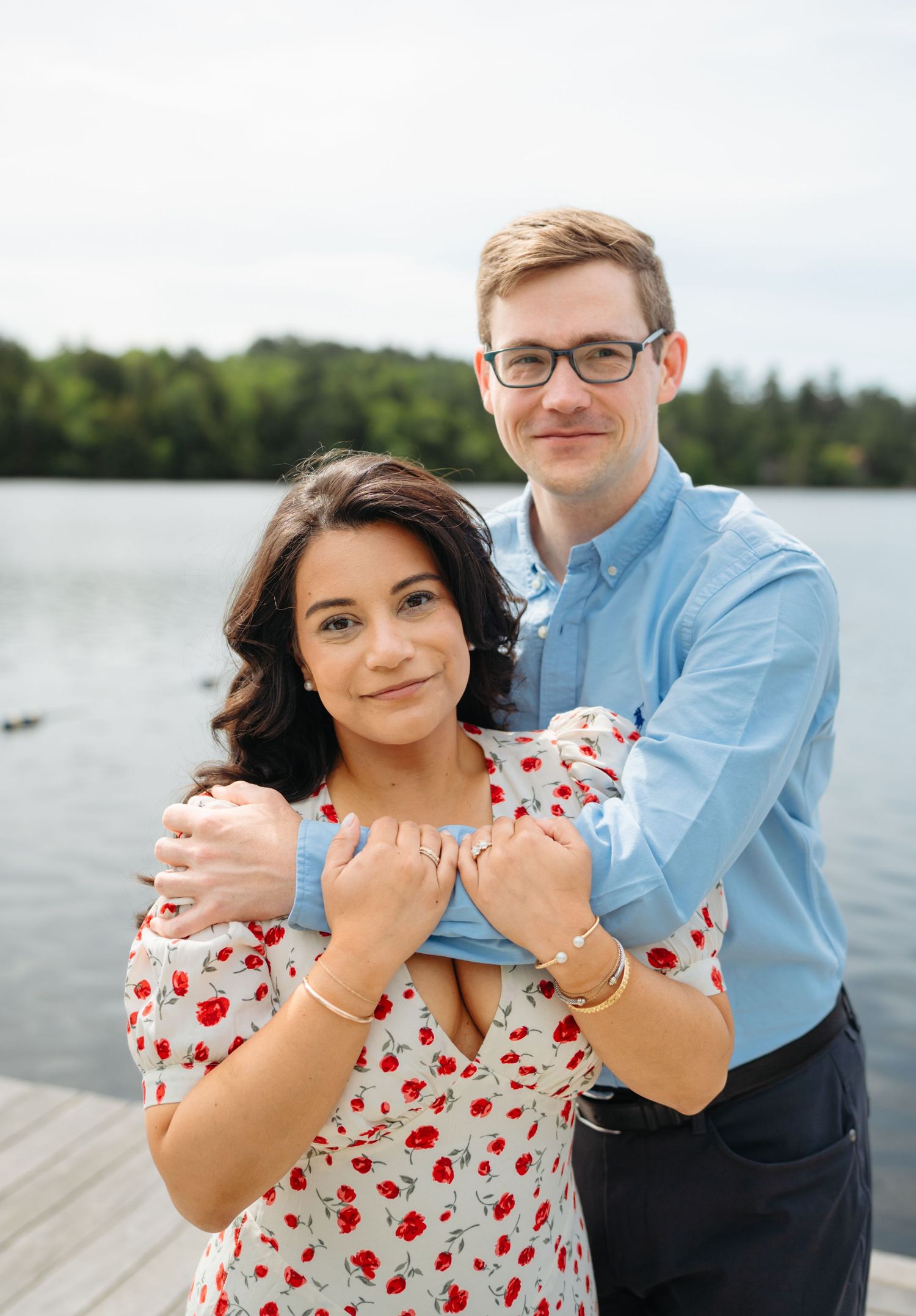 Couple posing on a lakeside dock, woman in floral dress, man in blue shirt, embracing with trees in background.