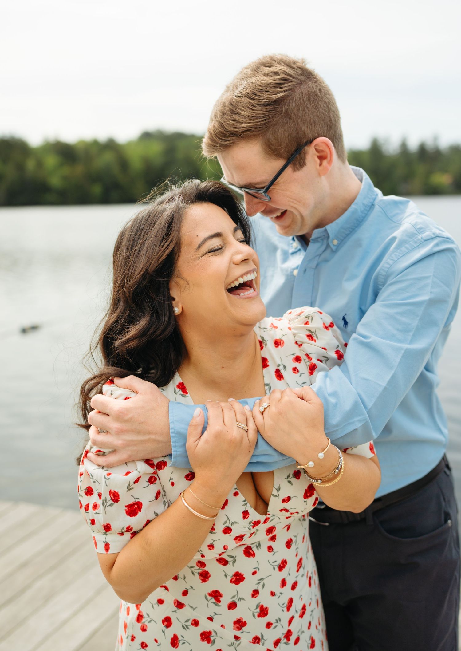 Laughing couple embracing on a lakeside dock, woman in floral dress and man in light blue shirt.