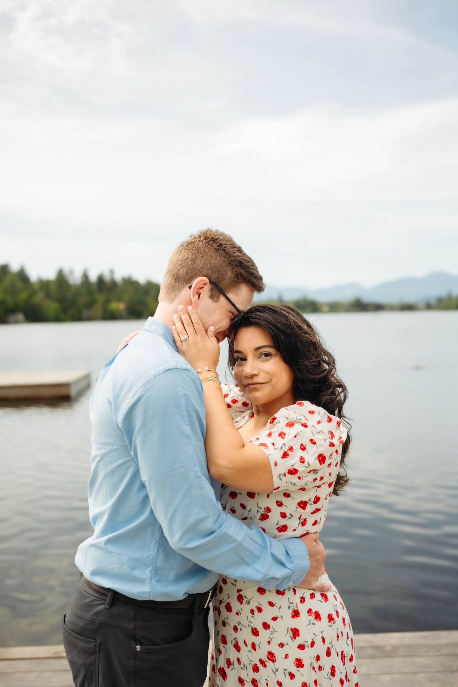 Romantic couple embracing on lakeside dock, woman in floral dress smiling, mountains visible in background.