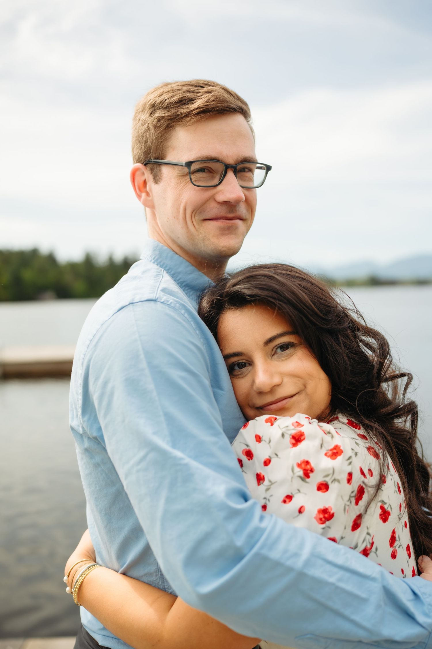 Couple hugging by a lake, man in glasses and blue shirt, woman in red floral dress smiling.