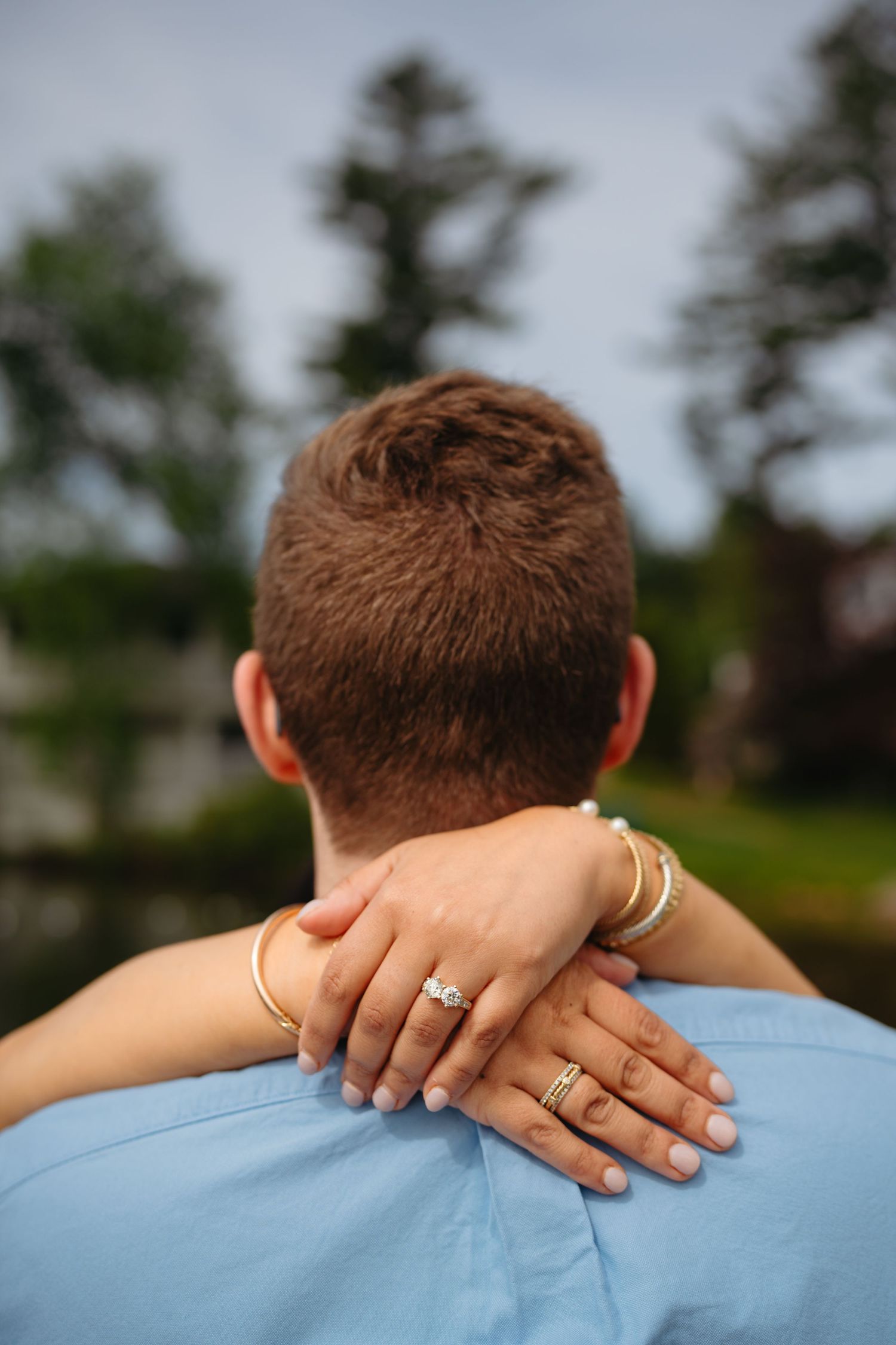 A woman's hands adorned with rings and gold bracelets rest on a man's shoulder from behind.
