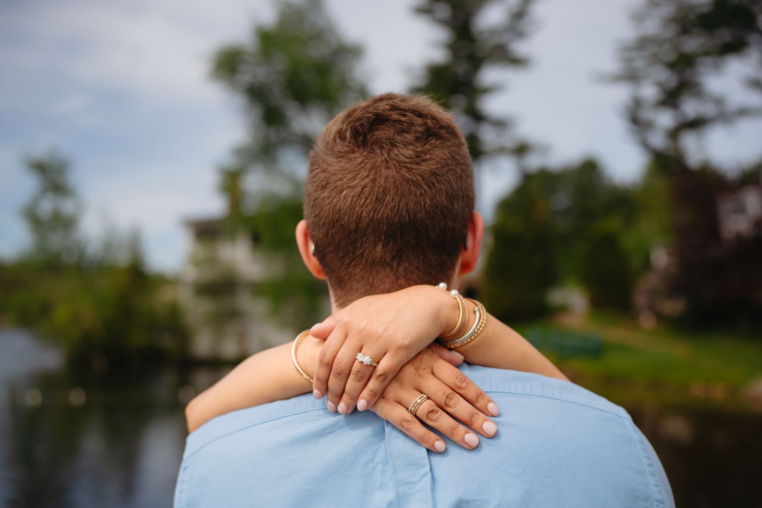 A couple embraces outdoors near a lake, woman's hand with rings and gold bracelet resting on man's shoulder.