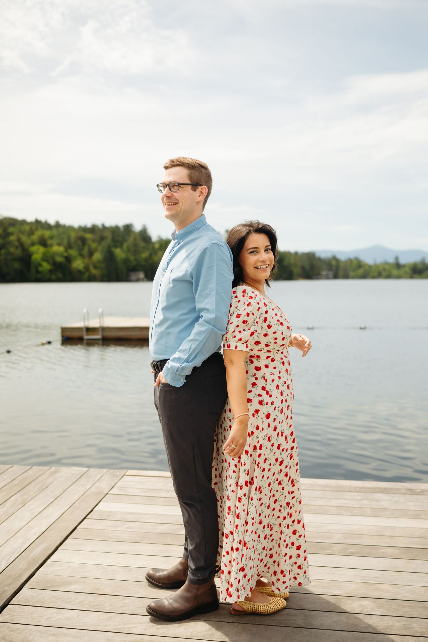 Couple standing back-to-back on a wooden dock by a lake, man in blue shirt and woman in floral dress, smiling.