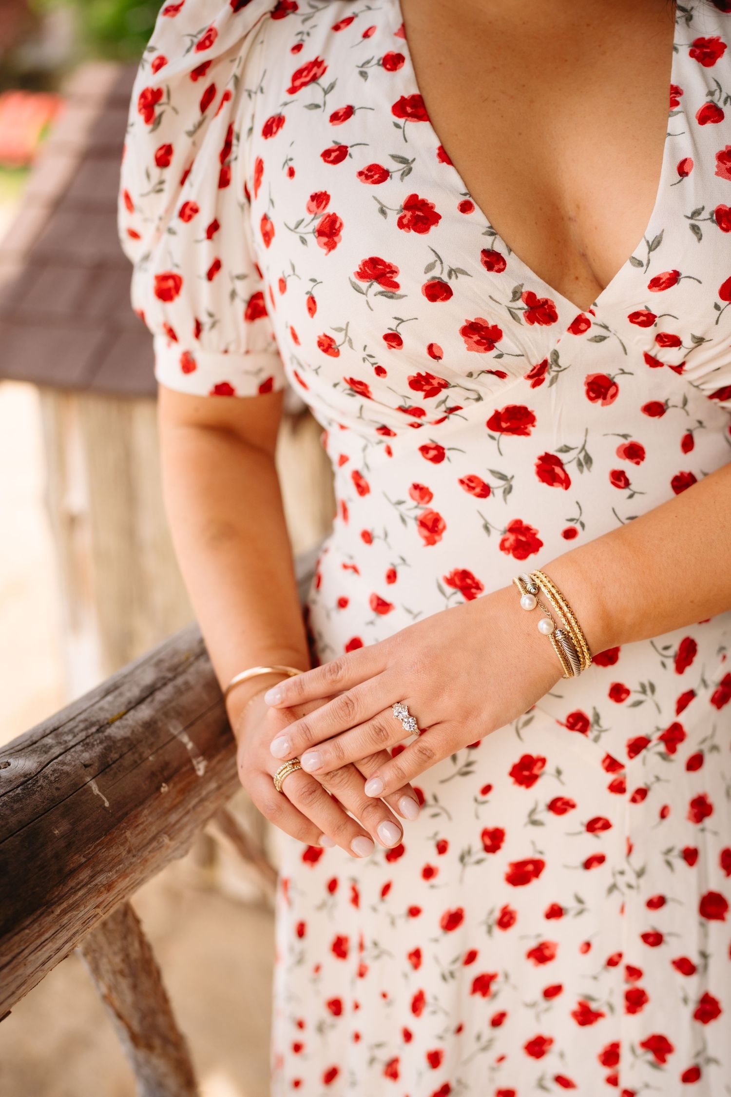 Woman in white floral dress with red roses, wearing gold bracelets and rings, resting hand on wooden railing.