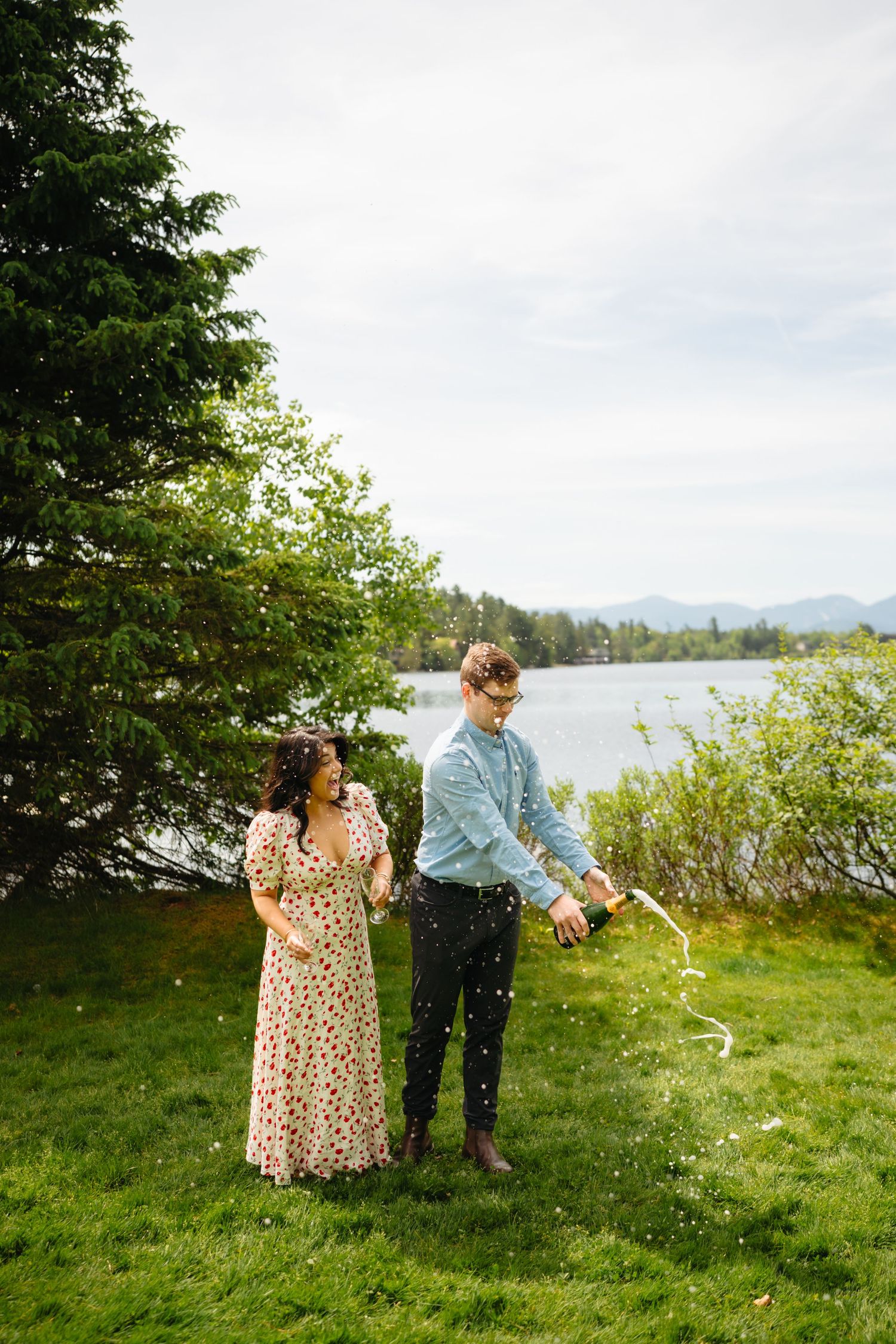 Couple celebrating outdoors by a lake, man popping champagne bottle while woman in floral dress watches, surrounded by greenery.