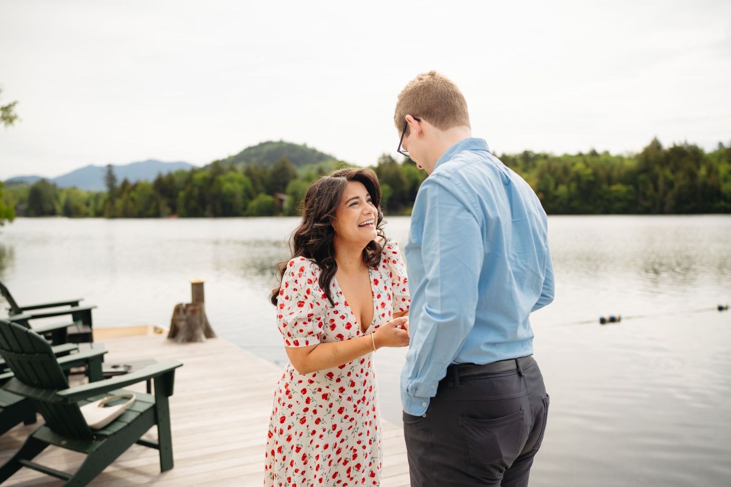 Couple laughing together on a lakeside dock, woman in floral dress, mountains in background.
