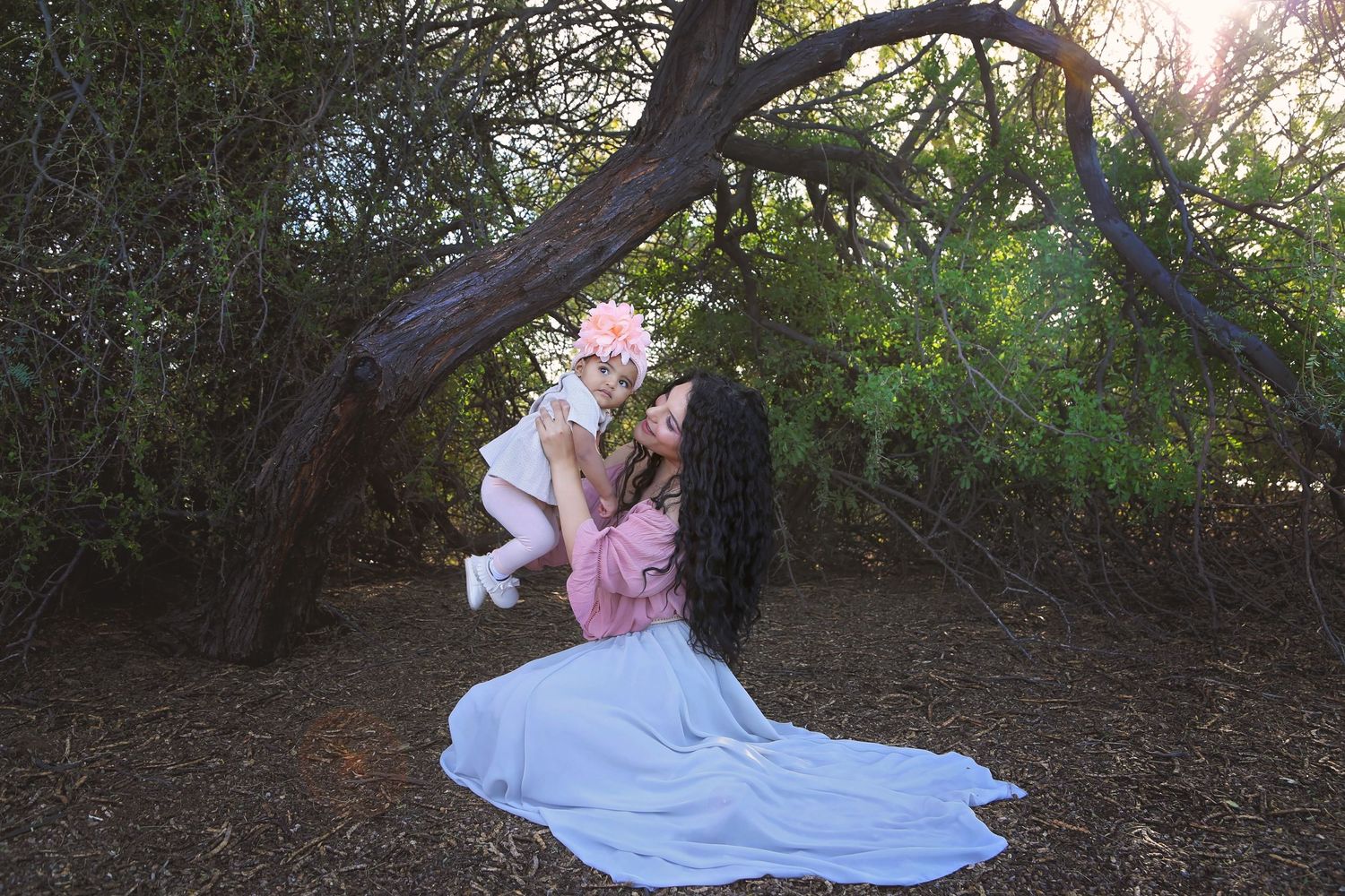 A mother in a flowing pale blue gown lifts her toddler wearing a pink flower crown beneath a twisting tree in a sunlit forest.