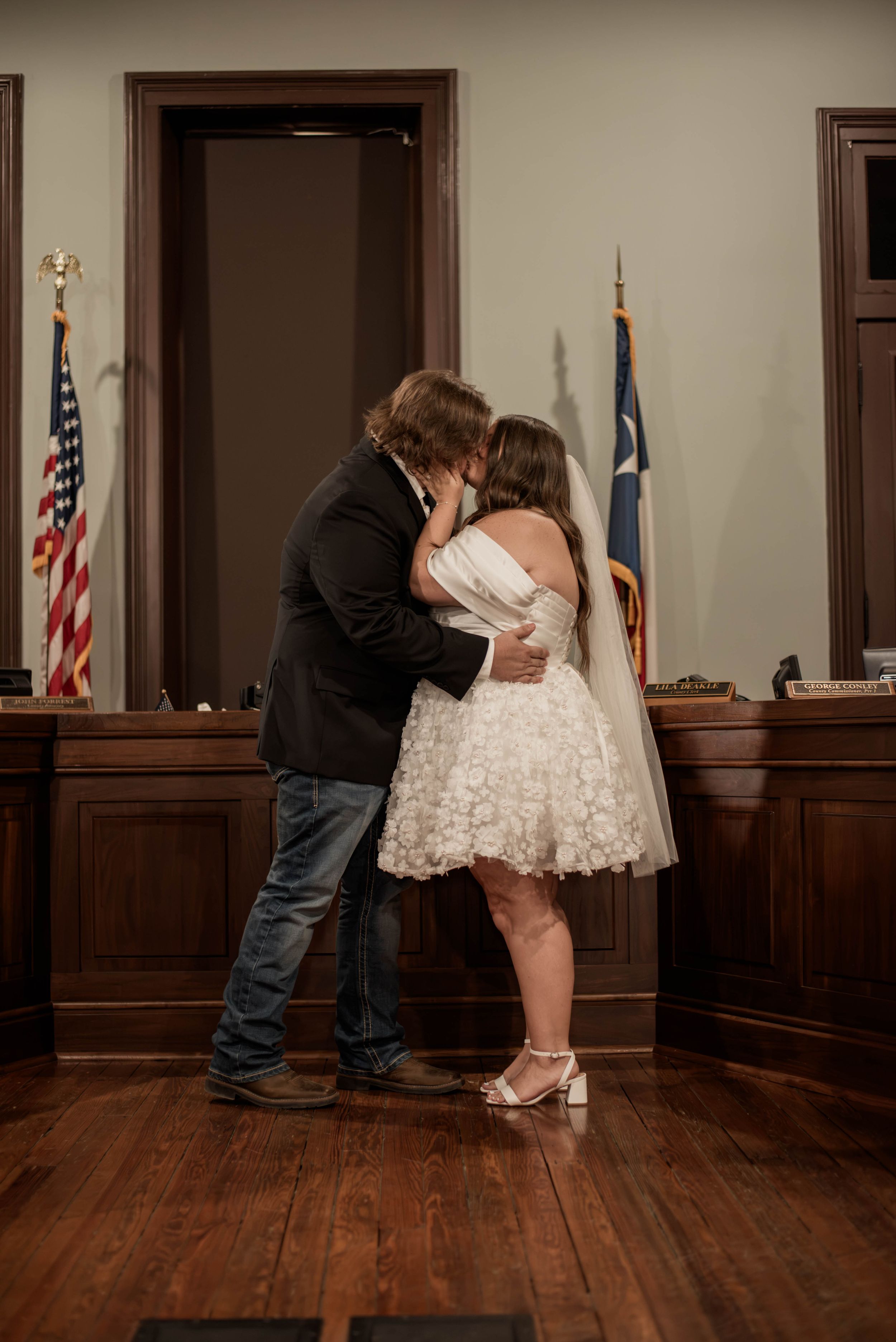 Newlyweds share their first kiss during a Weatherford Courthouse ceremony with Texas and American flags behind them.