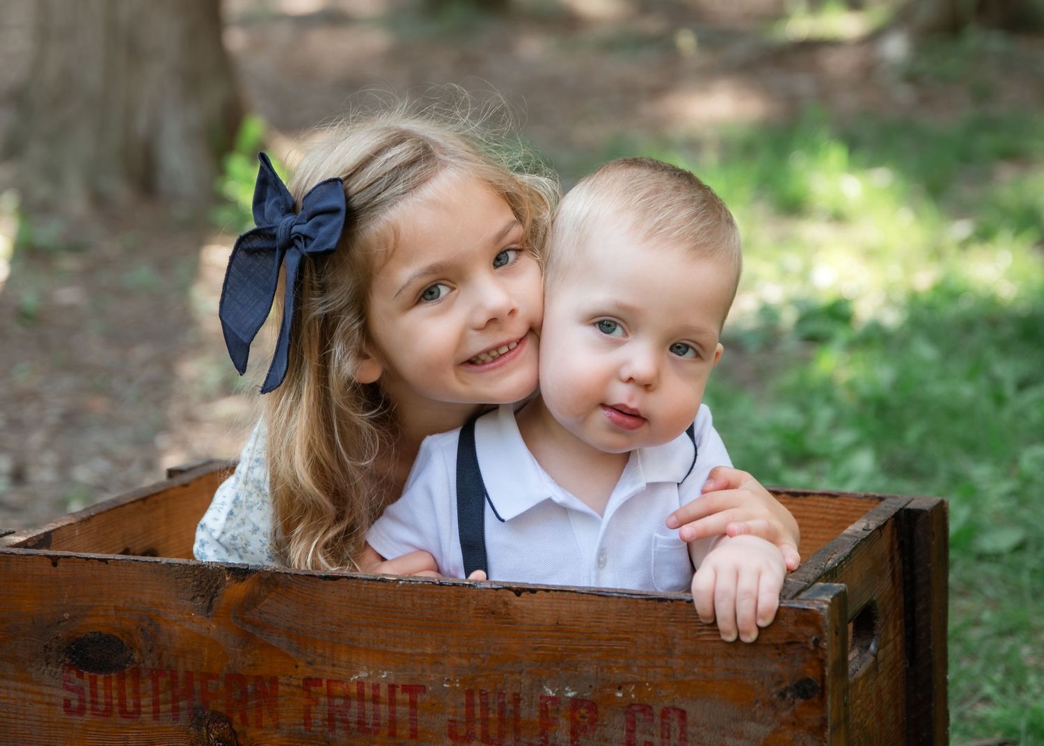 Two young siblings, a girl with a navy bow and a baby boy in suspenders, pose together in a rustic wooden crate outdoors.