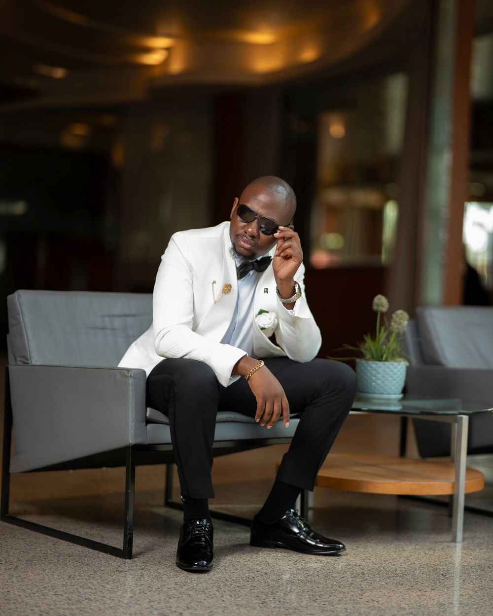 Stylish man in white blazer and bow tie sitting on gray sofa, adjusting sunglasses in modern lobby.