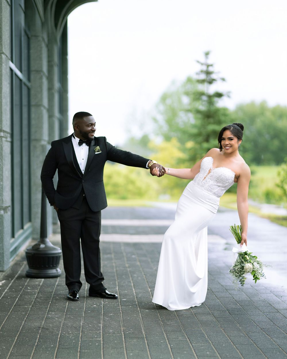 A groom in a black tuxedo holds his bride's hand as she walks away in a white gown holding a bouquet outdoors.
