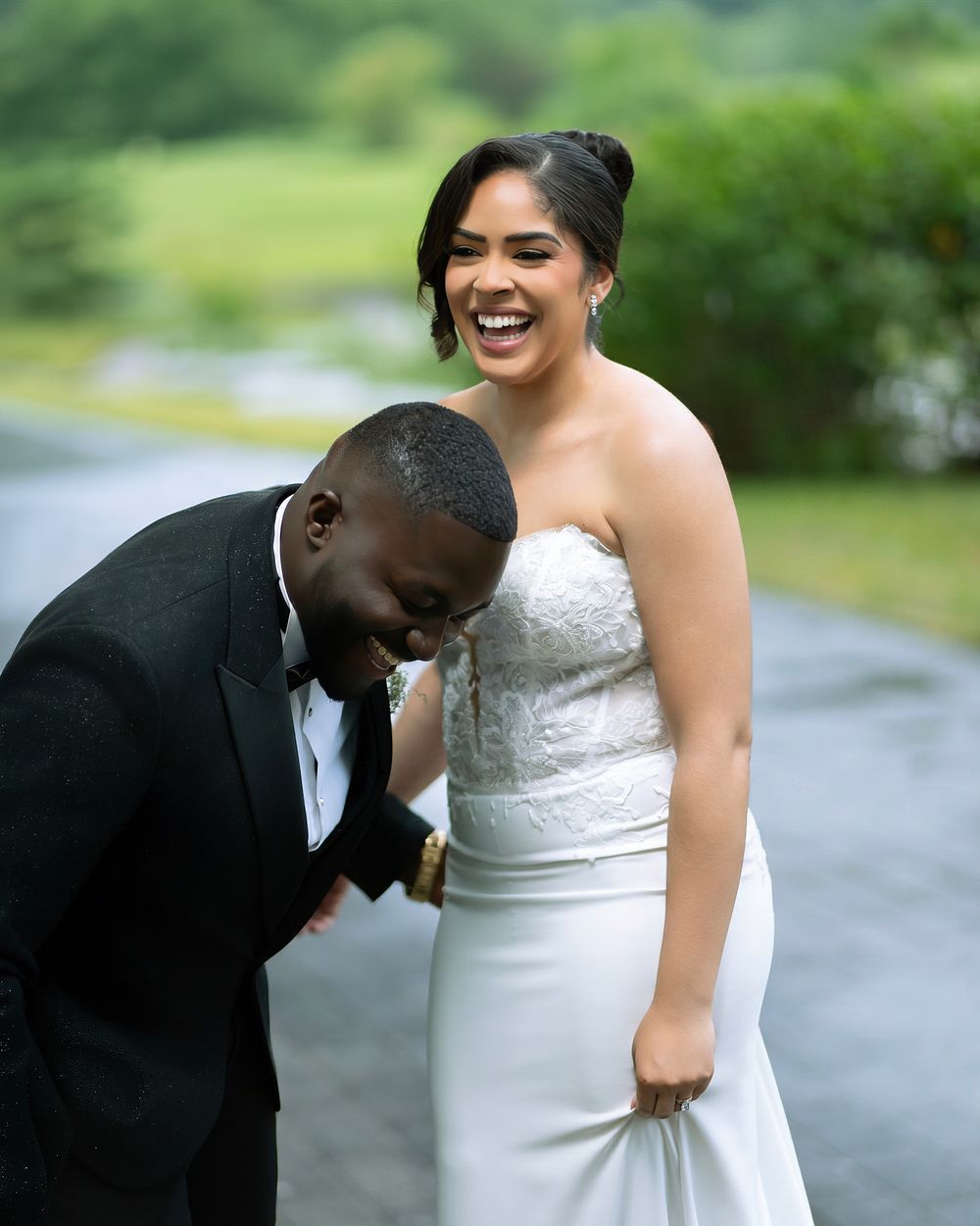 Joyful bride in strapless white gown laughs with groom in black tuxedo on a scenic outdoor path.