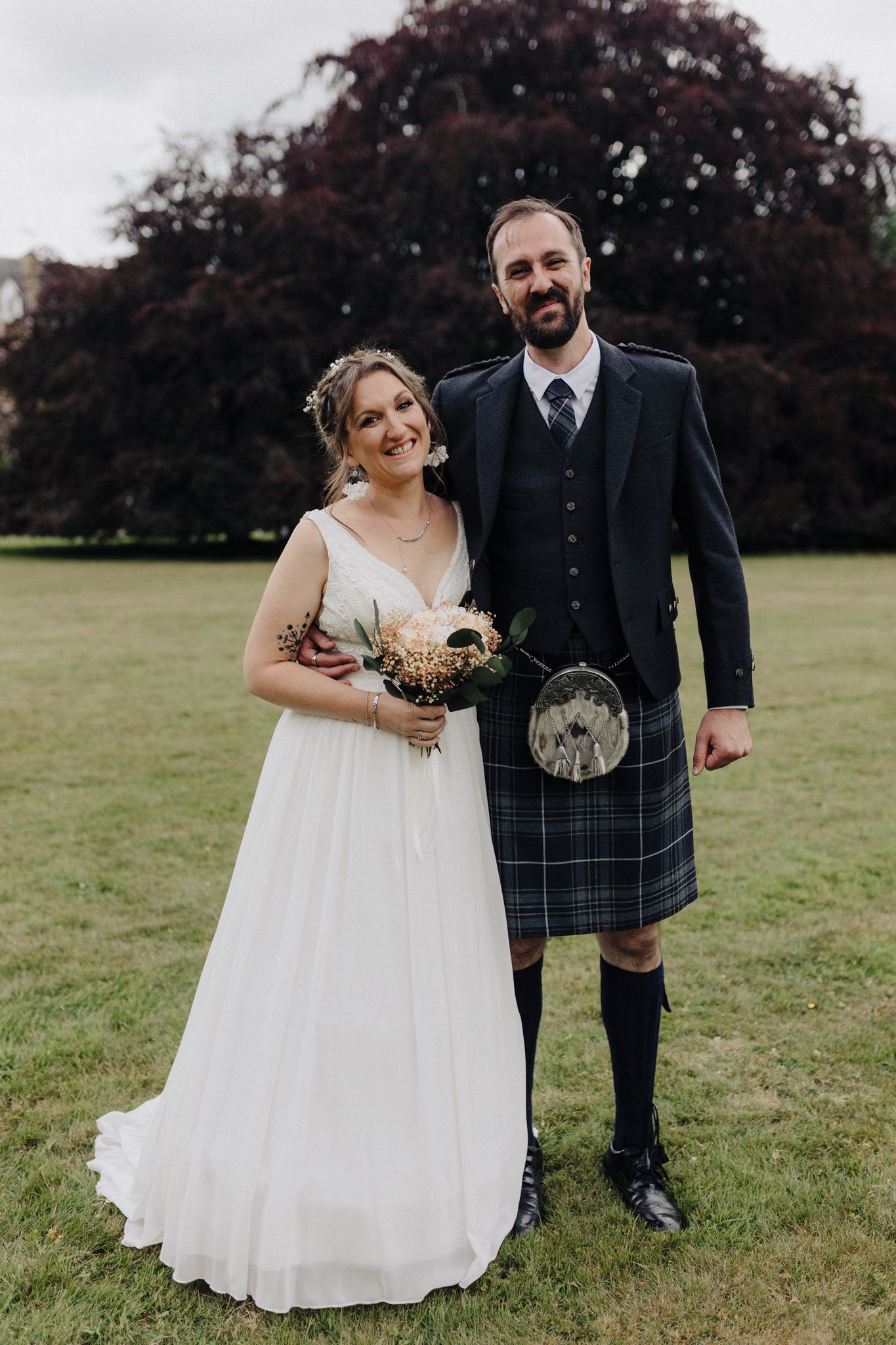 Bride in white gown and groom in Scottish tartan kilt pose together on green lawn.