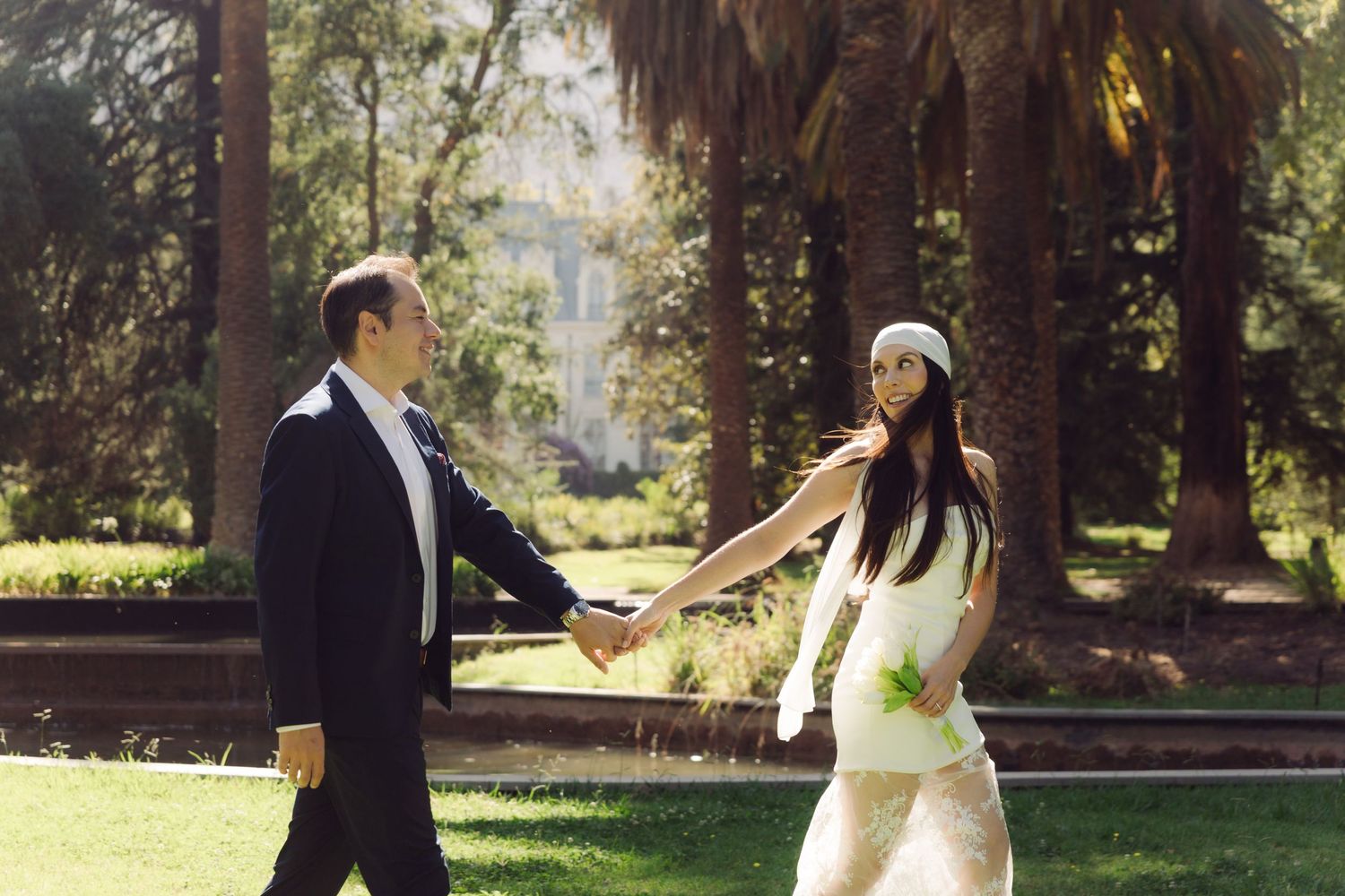 Bride and groom walk hand in hand through sunny park with tall palm trees in background.