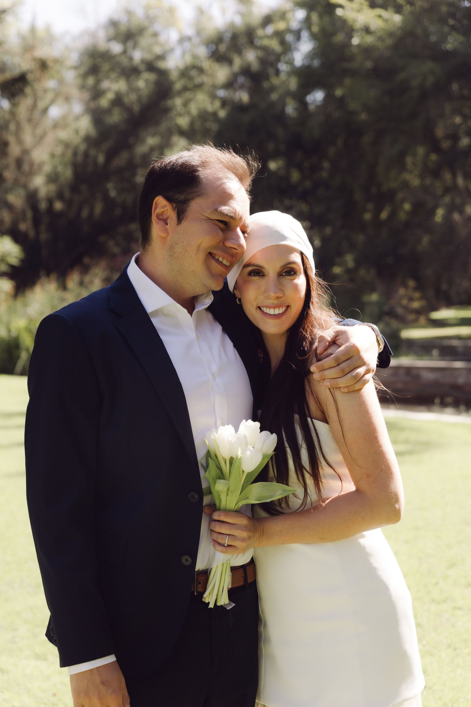 Smiling couple embraces outdoors, bride in white dress and headpiece holding white tulip bouquet.