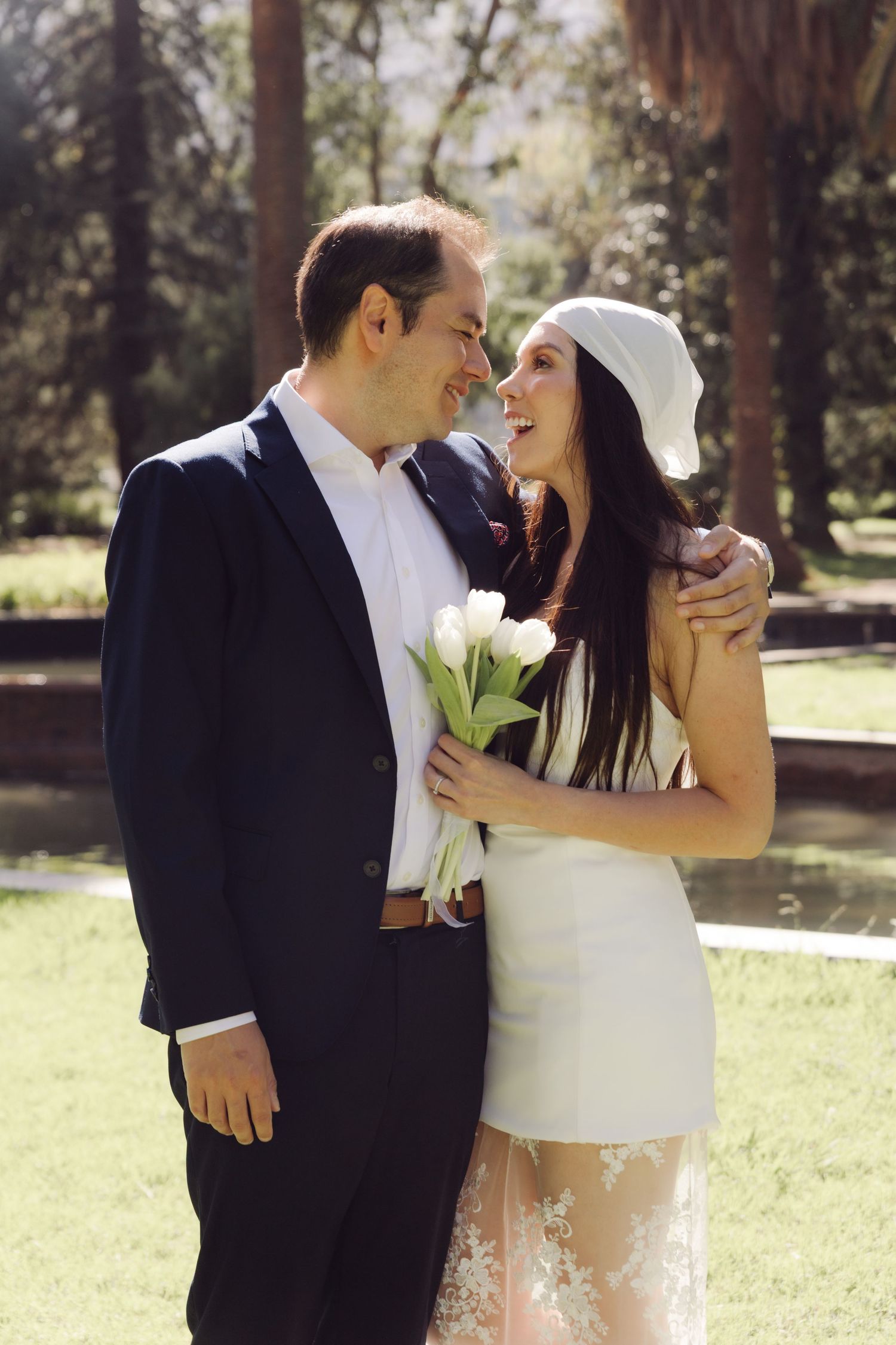 Smiling bride in white mini dress and headband holds tulips while groom in navy suit embraces her outdoors.