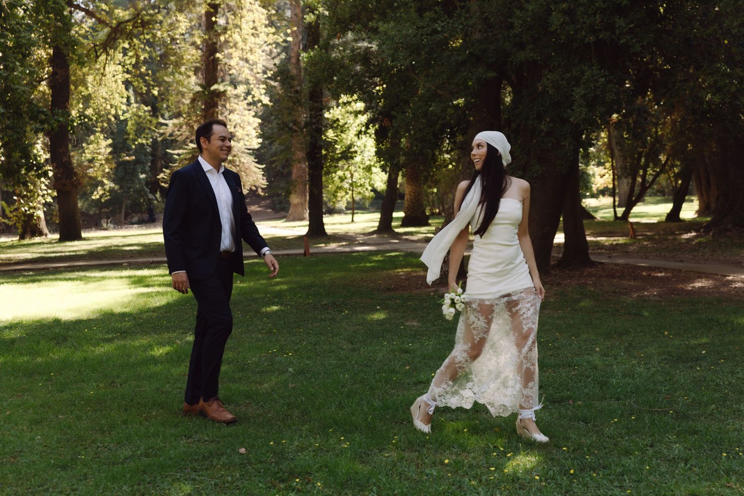 Bride in white lace dress and headband walks toward groom in navy suit on green lawn in park.