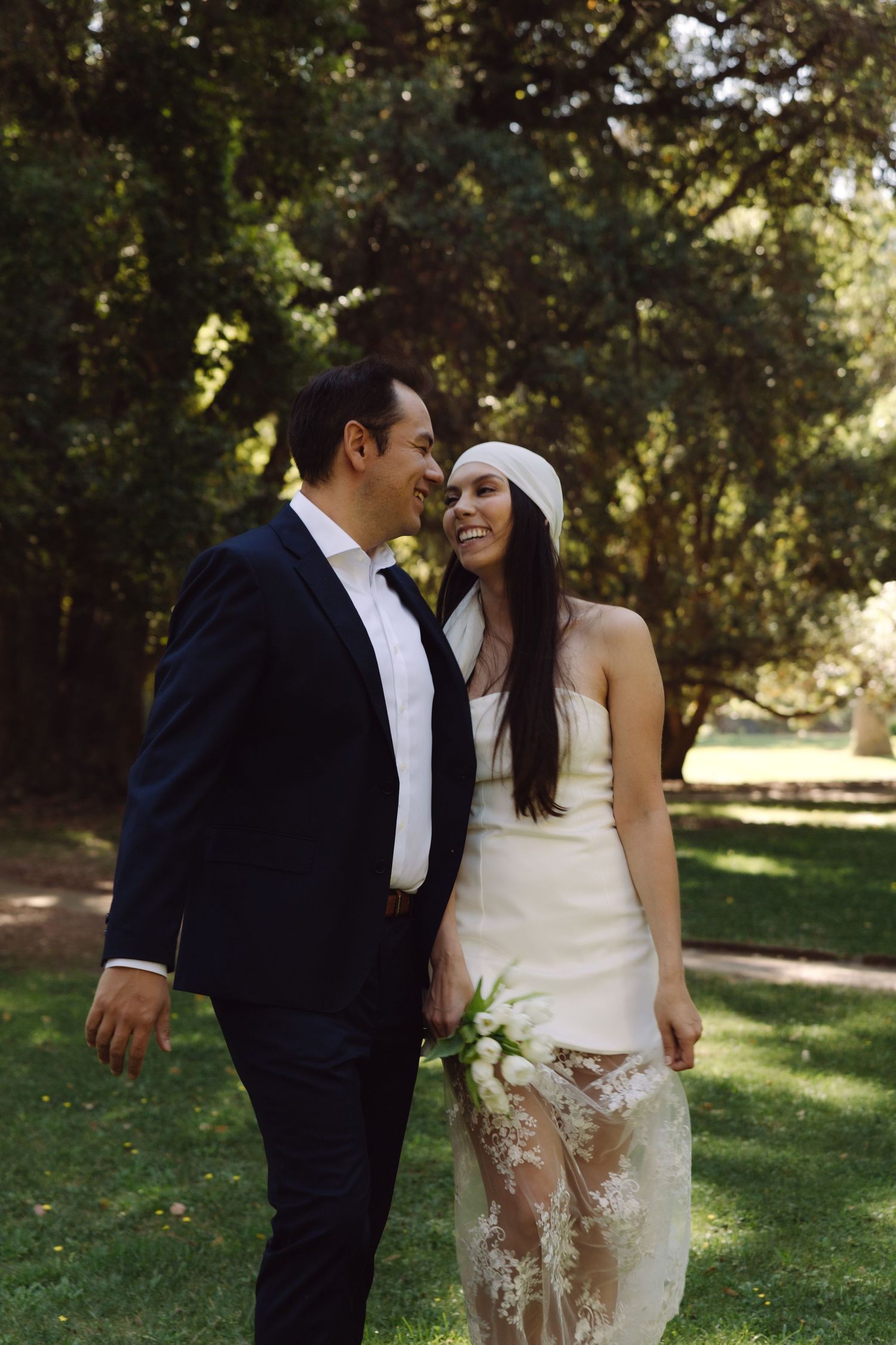 Happy bride and groom walk together in park, she wears white dress with lace overlay and white headband.