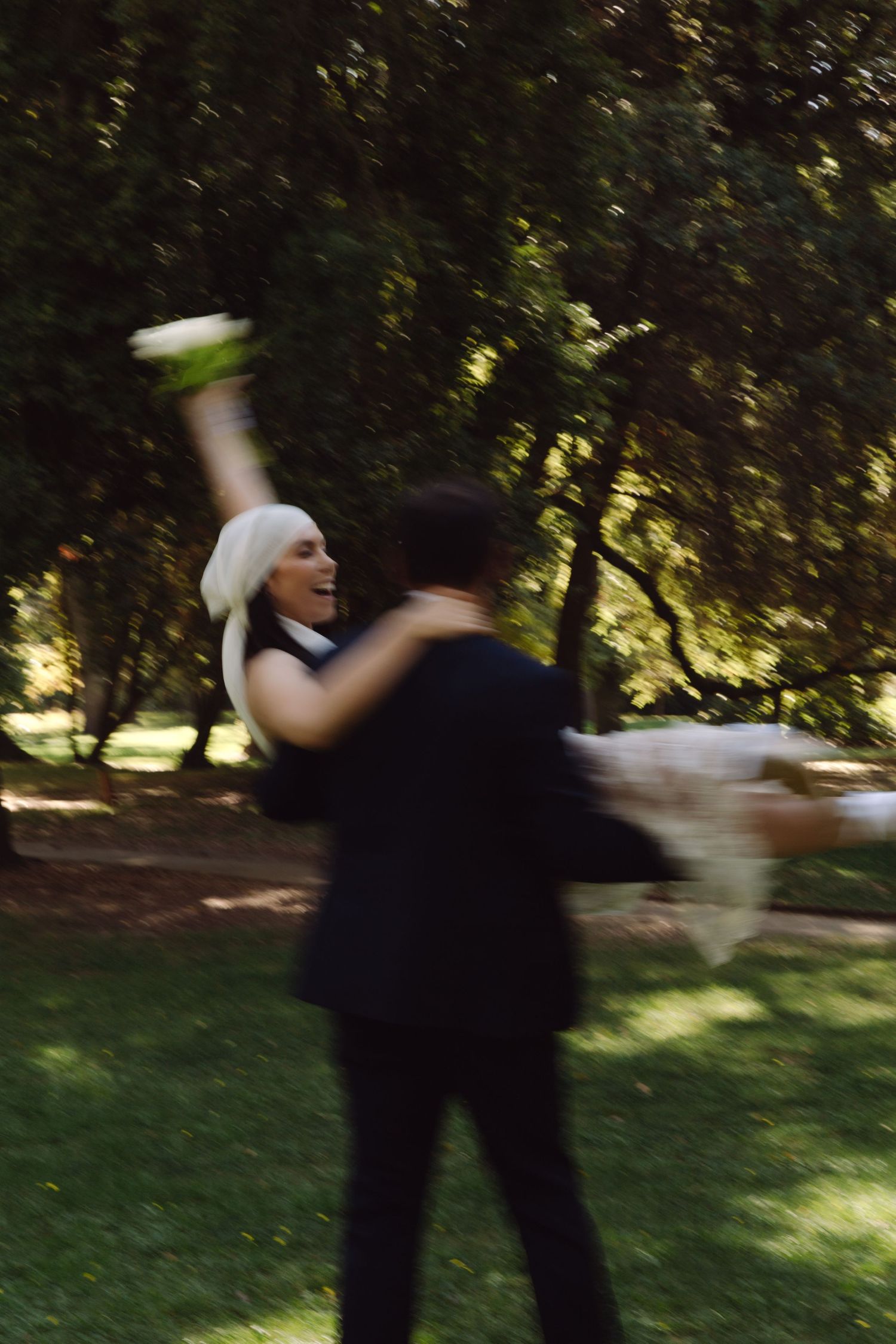 Blurred motion shot of groom lifting joyful bride in white dress with bouquet raised in park.