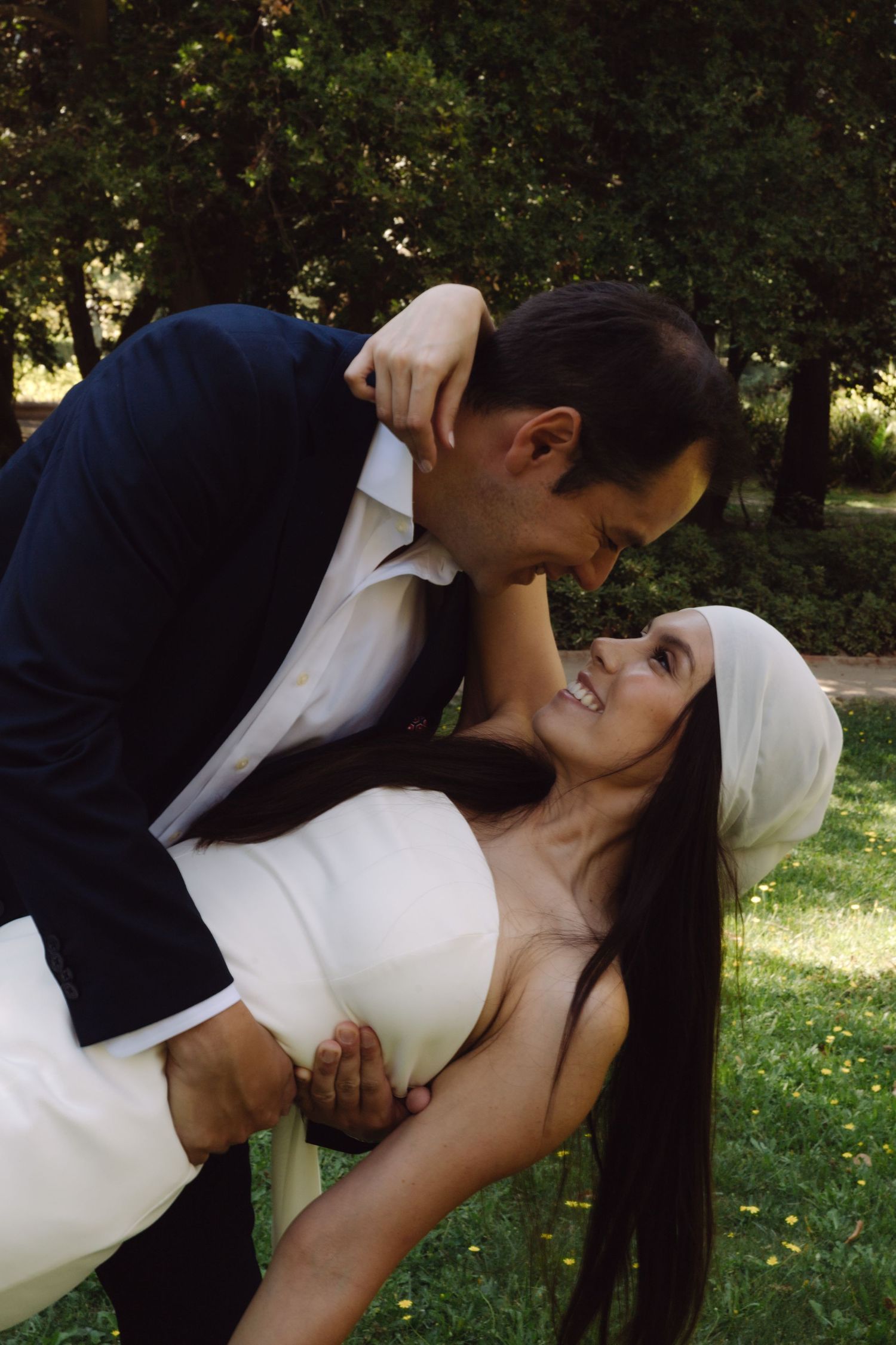 Groom in navy suit dips smiling bride in white strapless dress and white headband in lush garden.