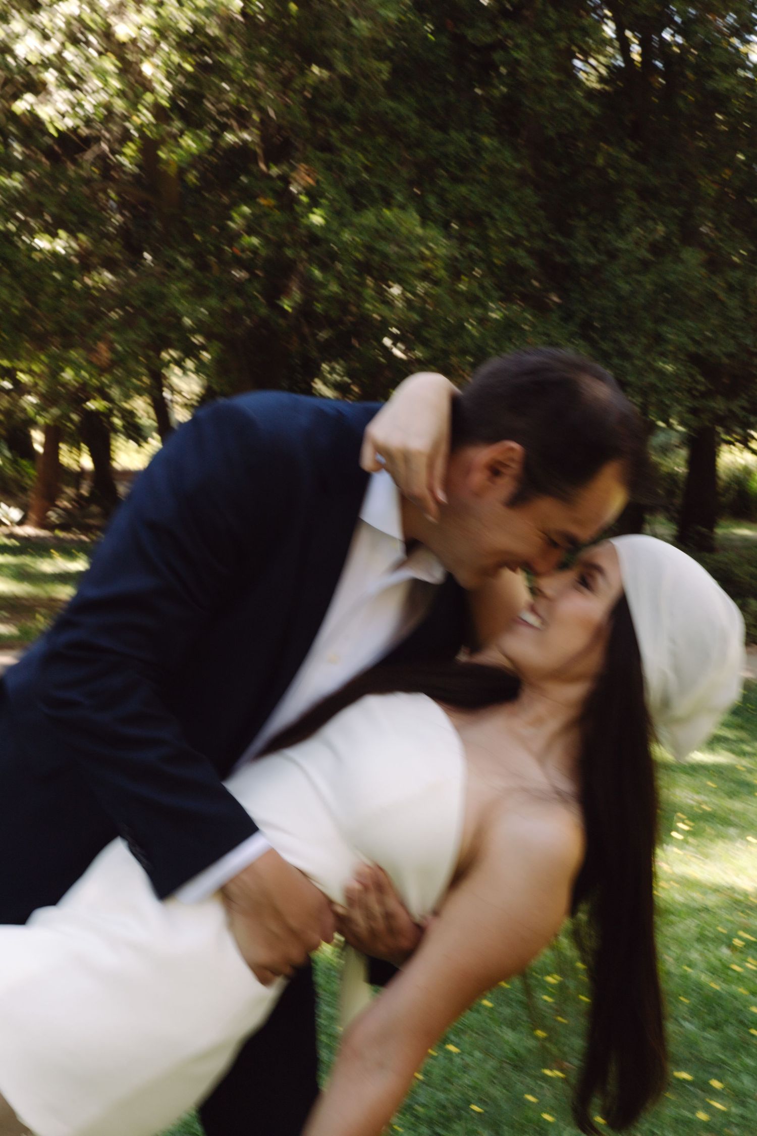 Groom leans in to kiss bride wearing white headband as he dips her in sun-dappled garden setting.