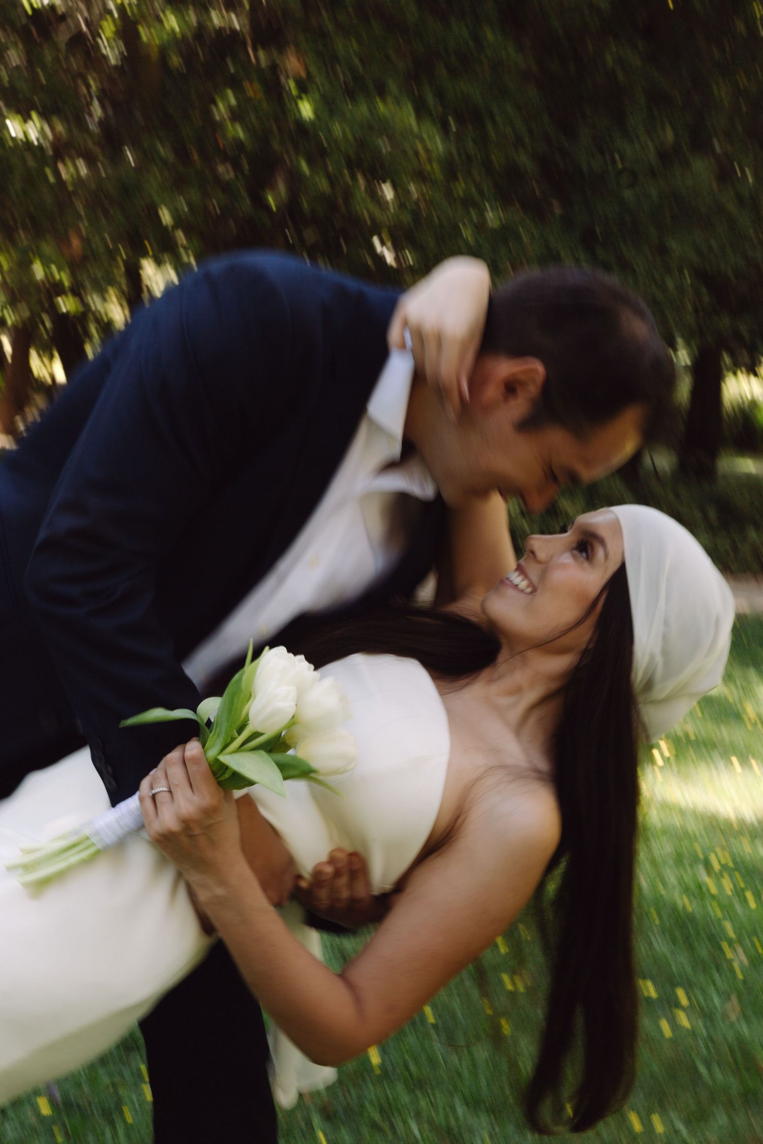 Groom dips bride holding white tulip bouquet for a romantic near-kiss in a green outdoor setting.