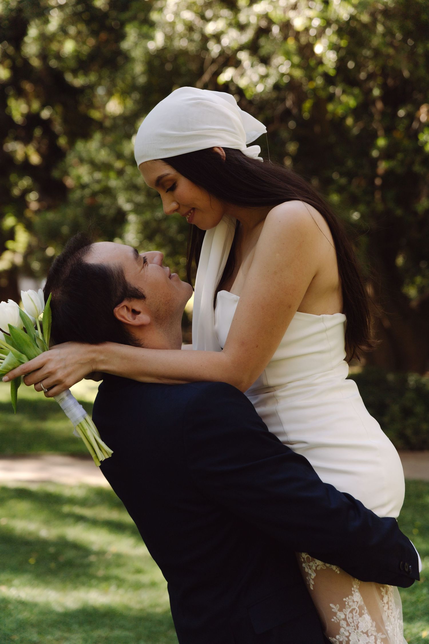 Groom lifts bride in white strapless dress and headband as they smile at each other in garden.