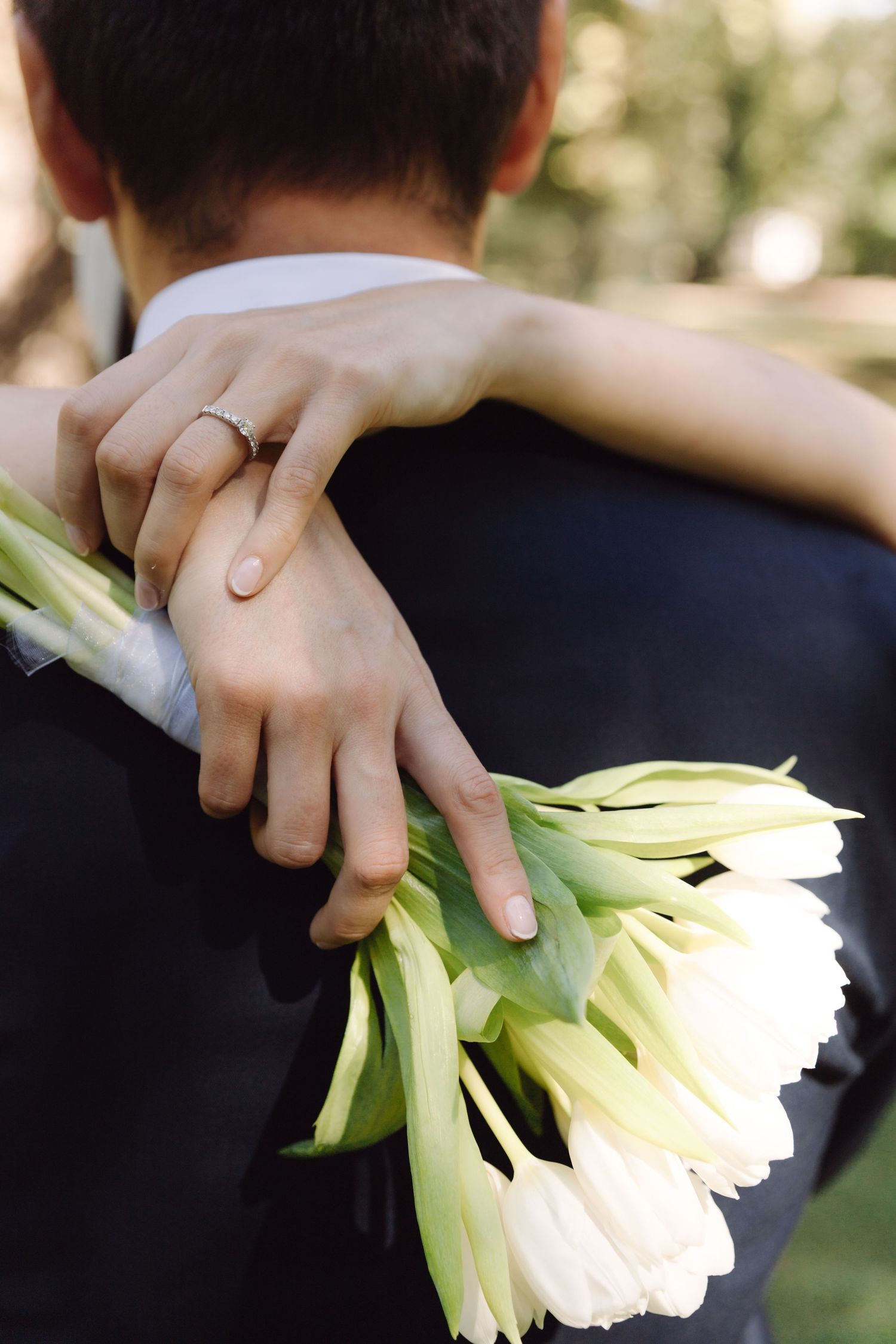 Close-up of bride\u2019s hands with diamond ring embracing groom\u2019s back while holding white tulip bouquet.
