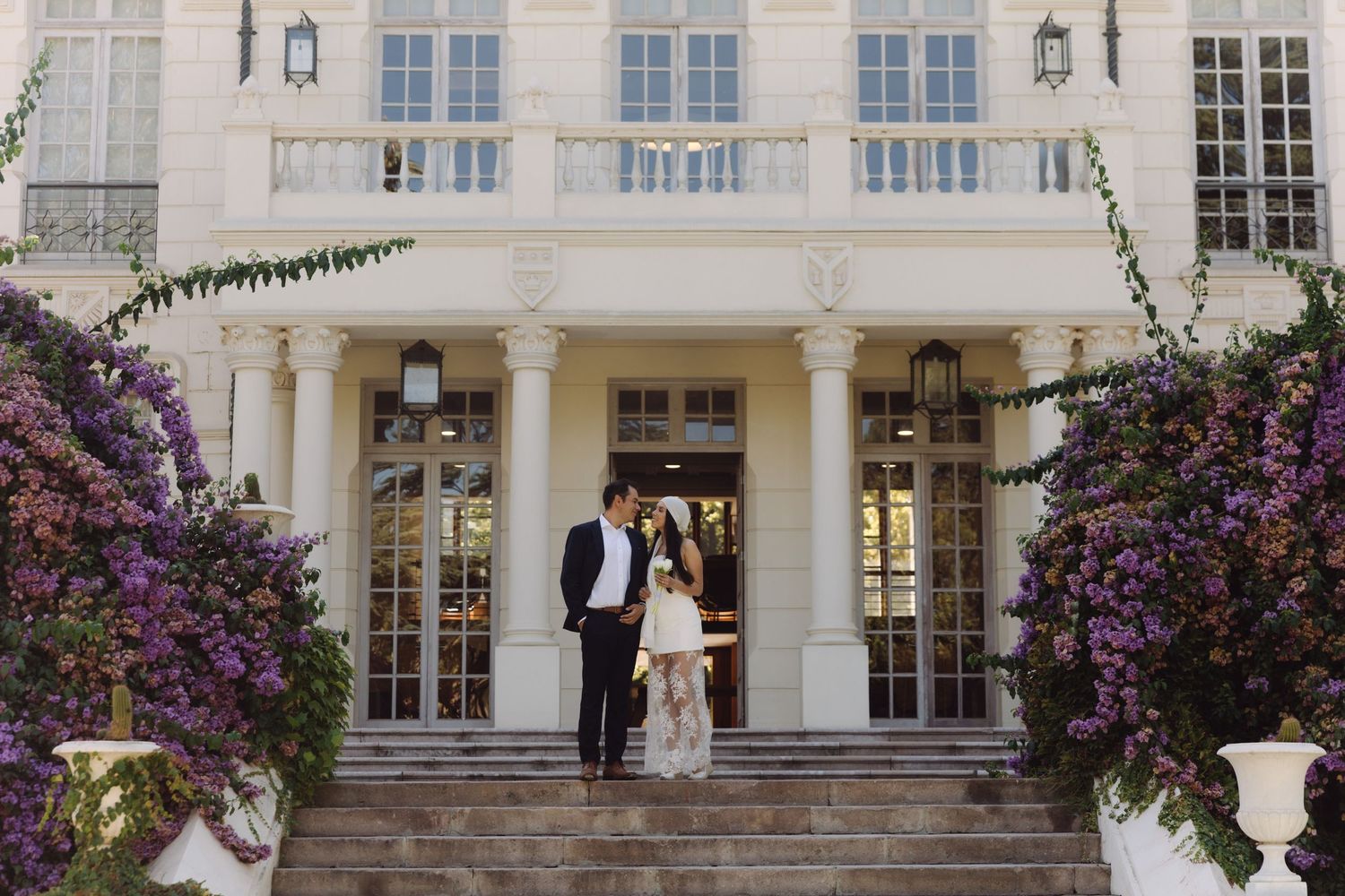 Newlyweds embracing at entrance of luxurious white estate framed by lush purple wisteria blooms.