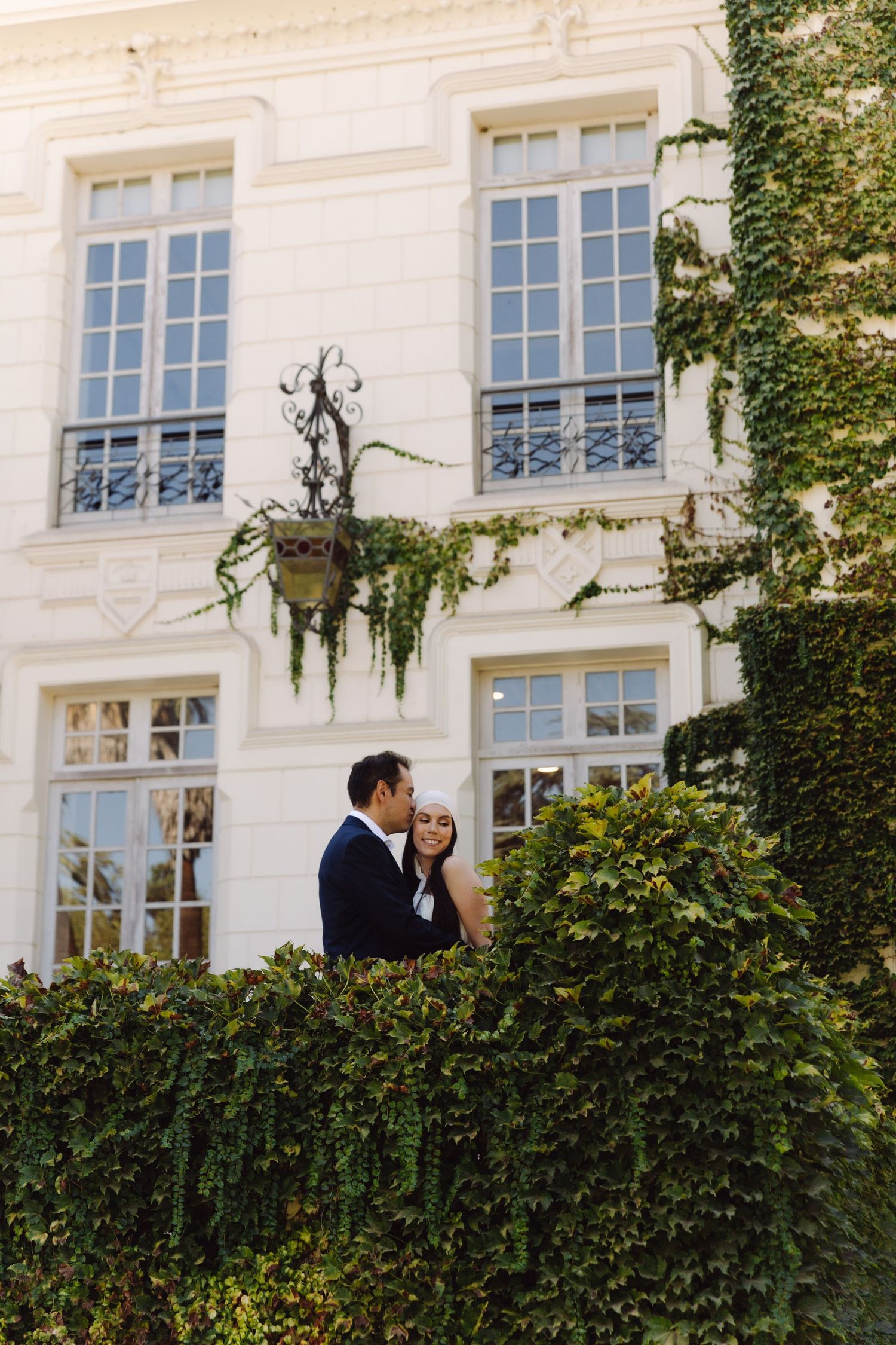 Romantic couple embracing behind ivy-covered white mansion wall in soft golden sunlight.